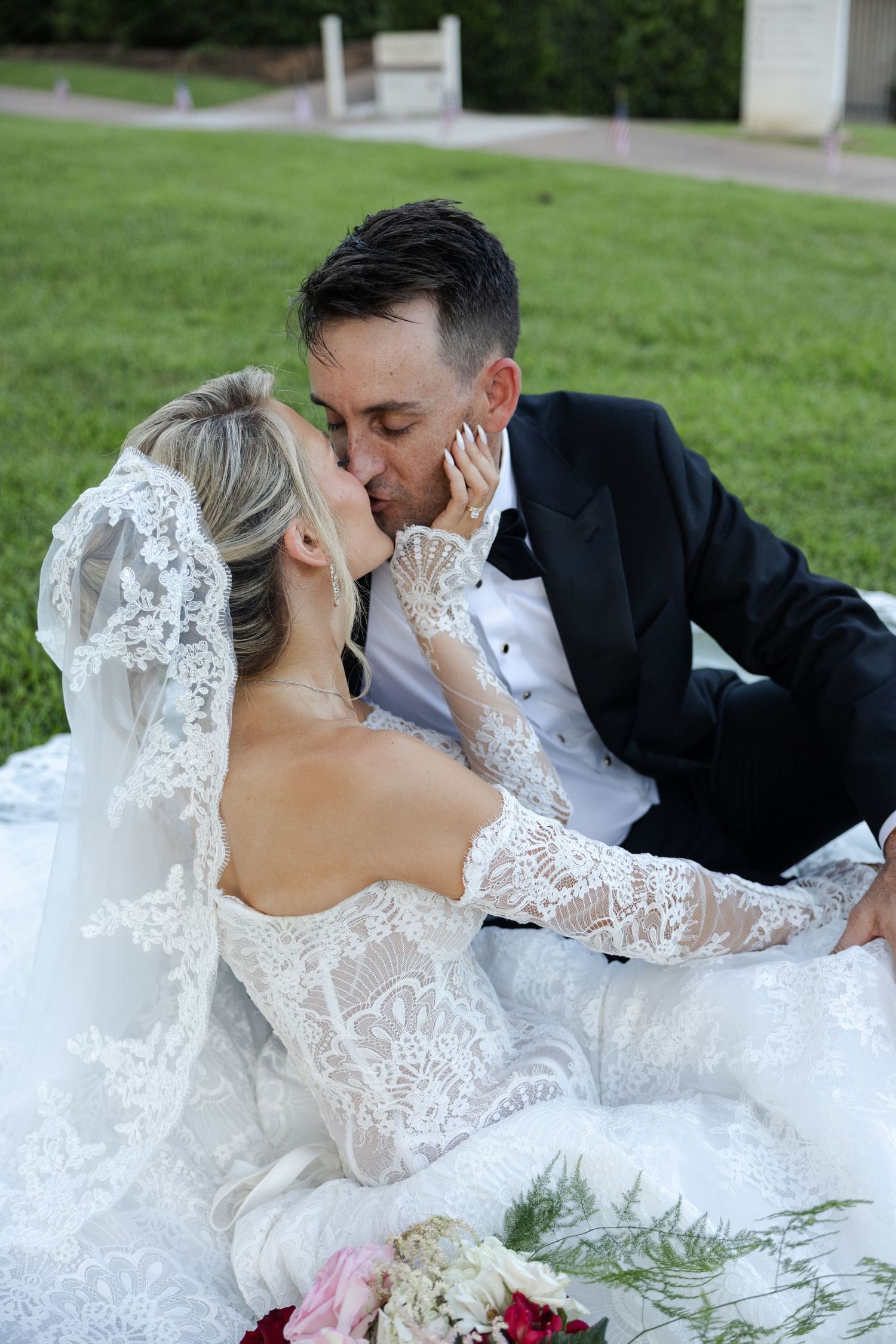 Bride and groom sharing a romantic kiss while sitting on green grass, bride wearing lace wedding gown with veil.