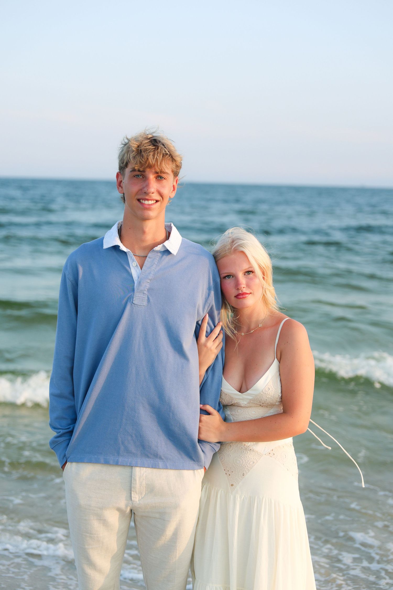 Young couple smiling together on a beach, man in blue polo and woman in white dress with ocean waves behind them.