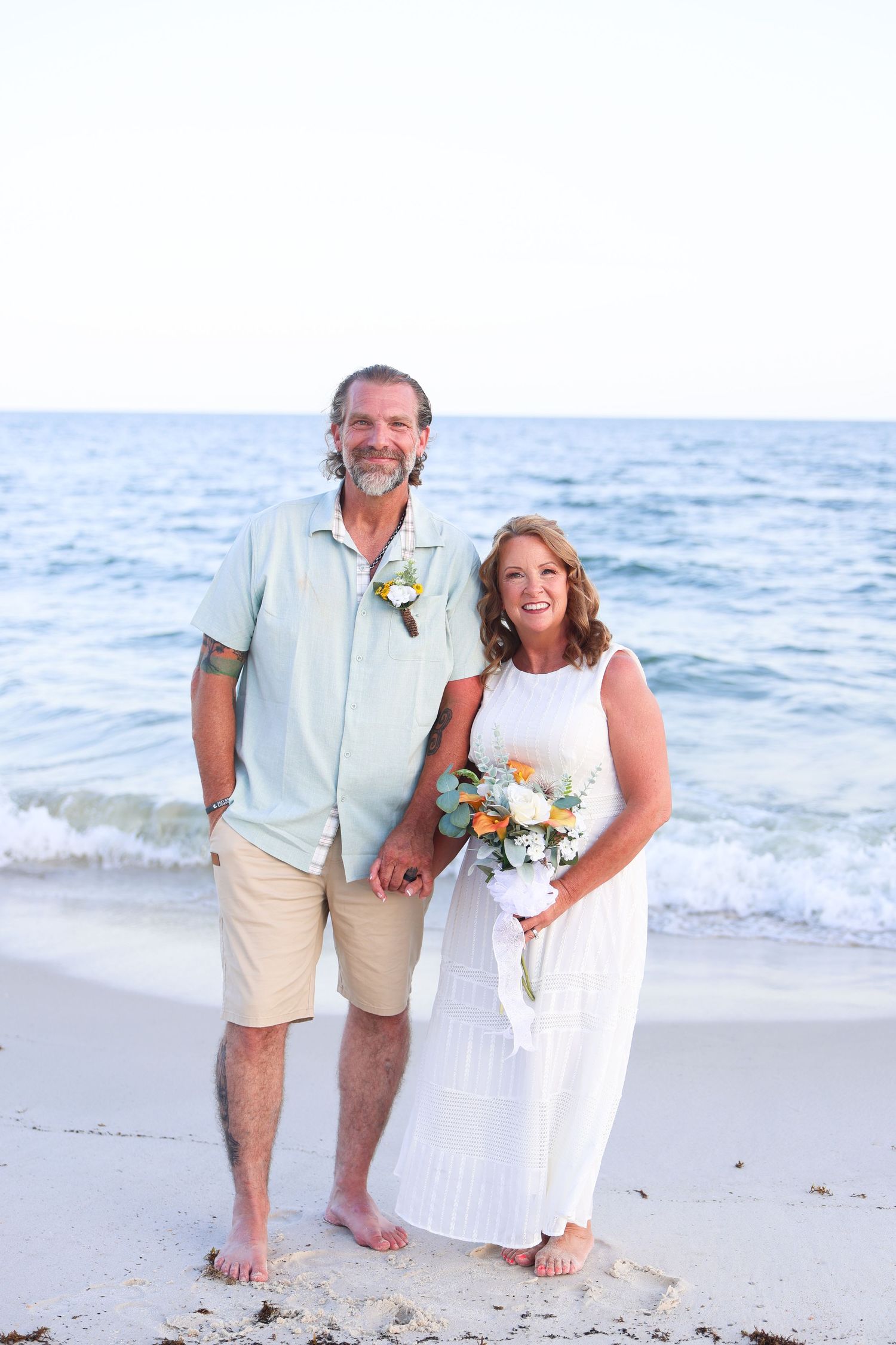 Couple in wedding attire holding hands and bouquet on a sunny beach with ocean waves behind them.