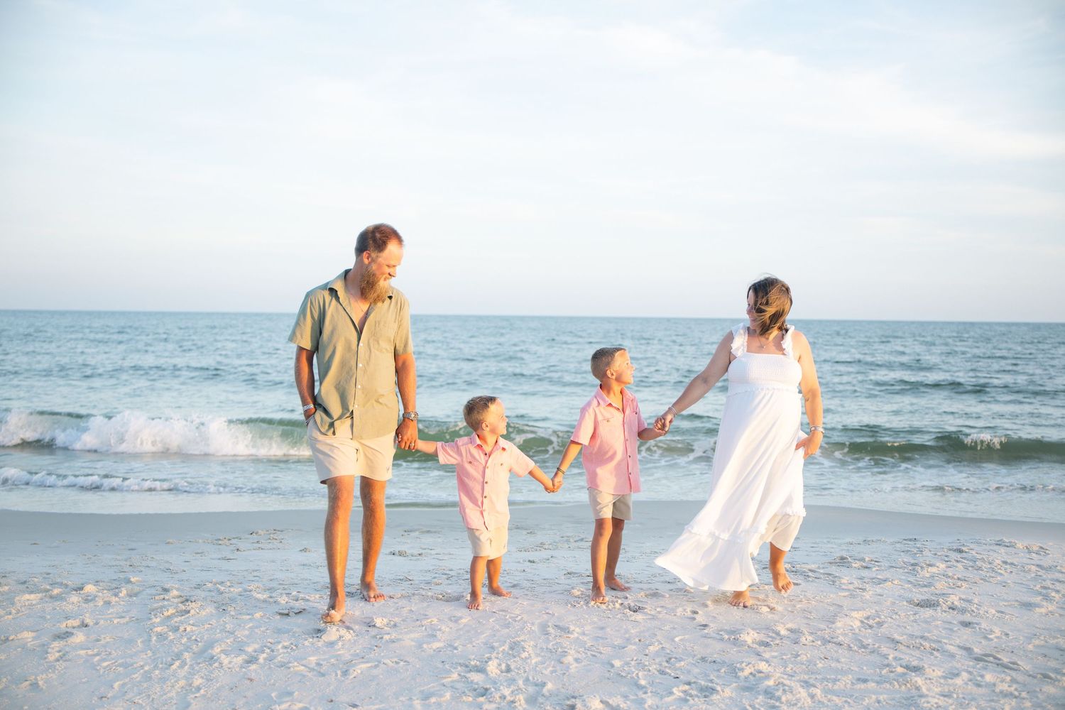 Family of four holding hands and walking on a sunny beach with gentle waves, wearing light pastel clothing.