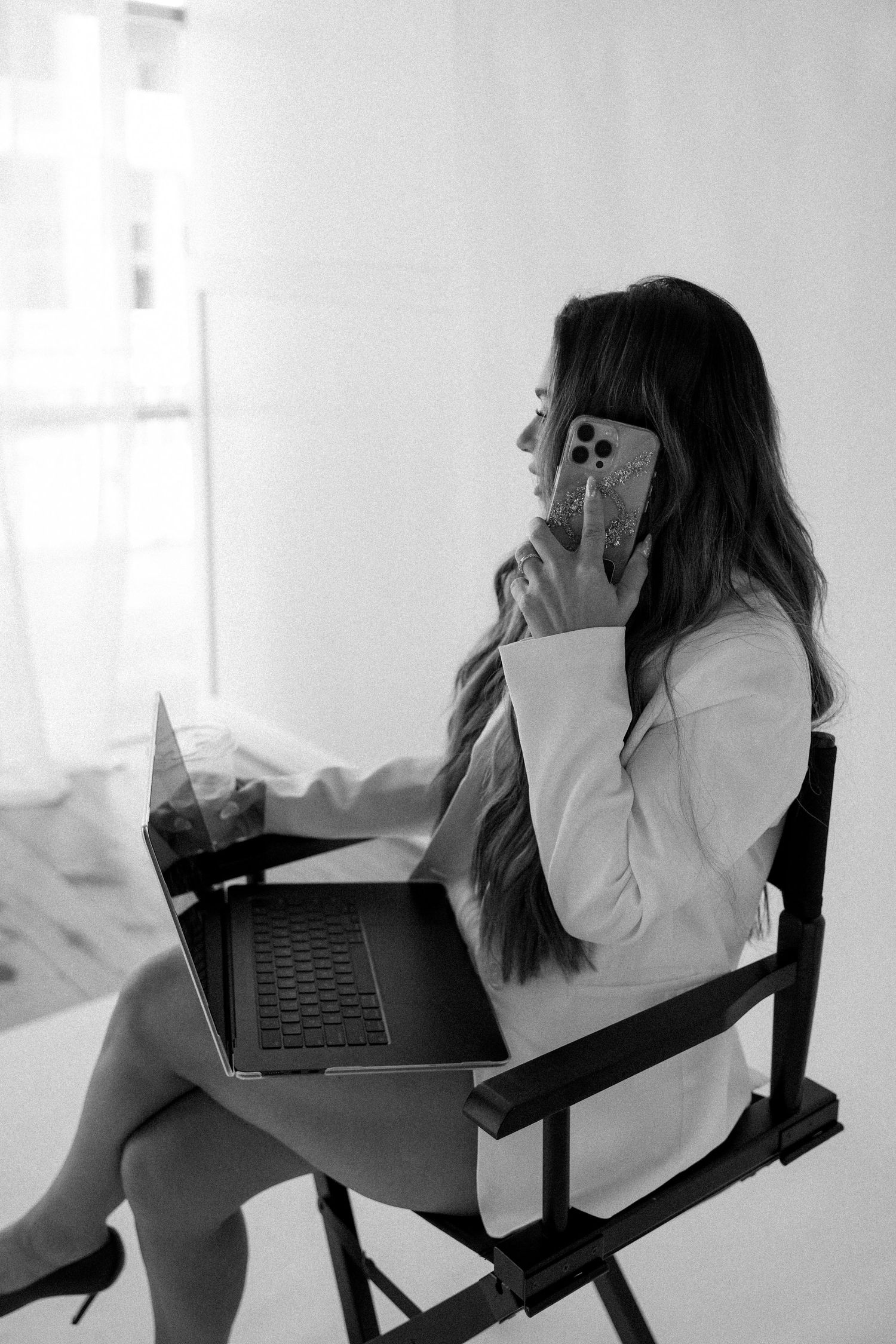 Professional woman in white blazer sitting on chair, using laptop and talking on phone in black and white photo.
