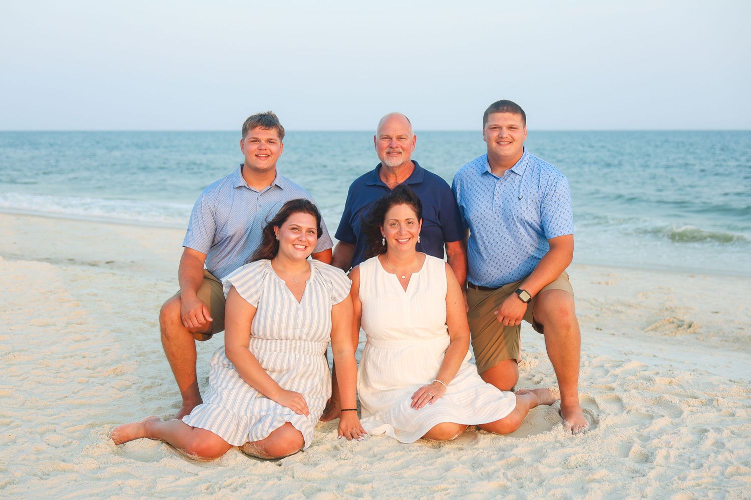 Family of five posing on a sandy beach at sunset, with two women seated and three men standing behind them.