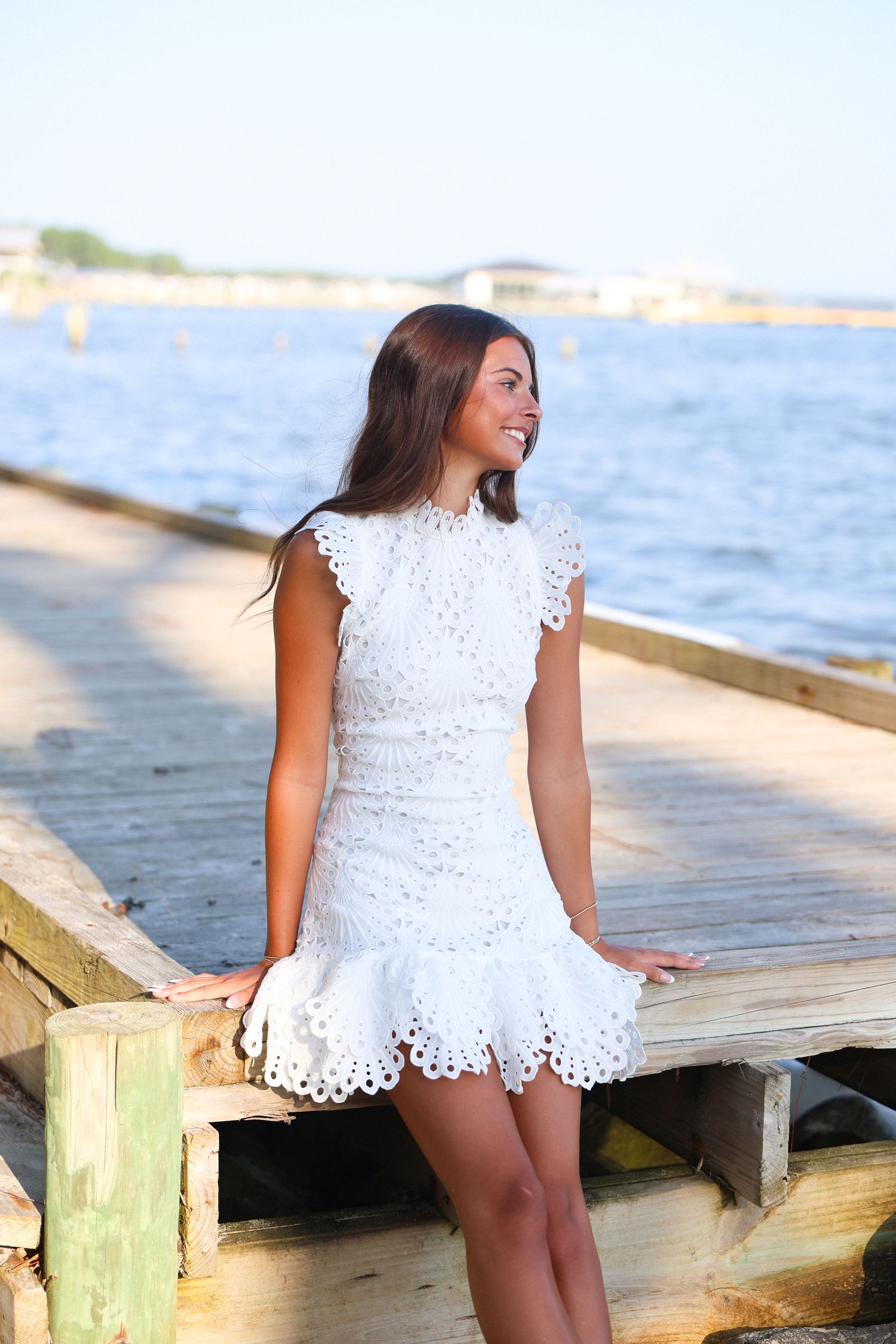 Young woman in white lace dress sitting on wooden pier by the water, smiling and looking to the side.