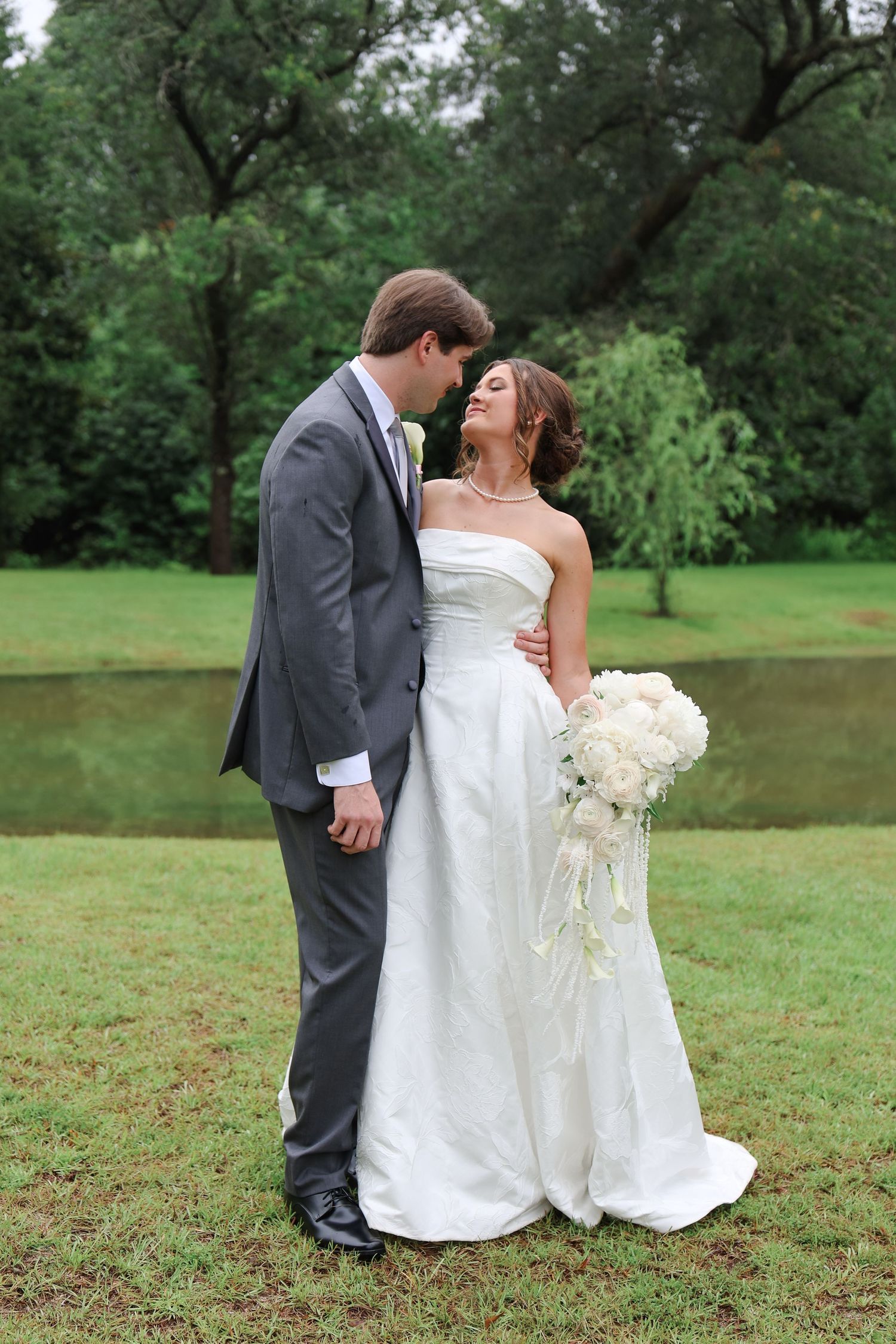 Bride in white strapless gown holding bouquet stands with groom in gray suit near pond surrounded by lush green trees.