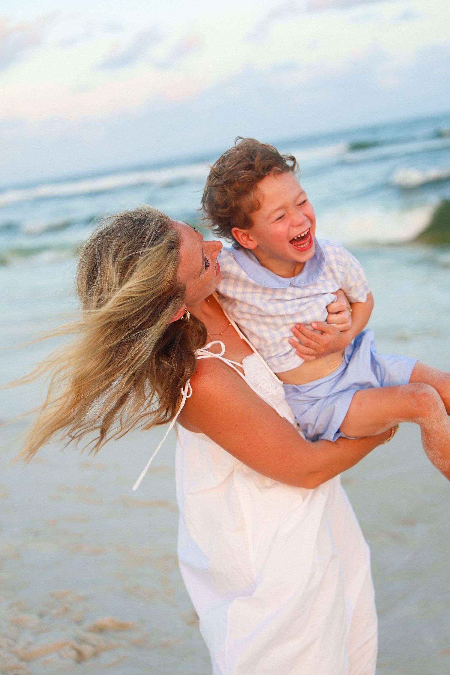 A joyful mother in a white dress spins her laughing young son on a beautiful sandy beach.
