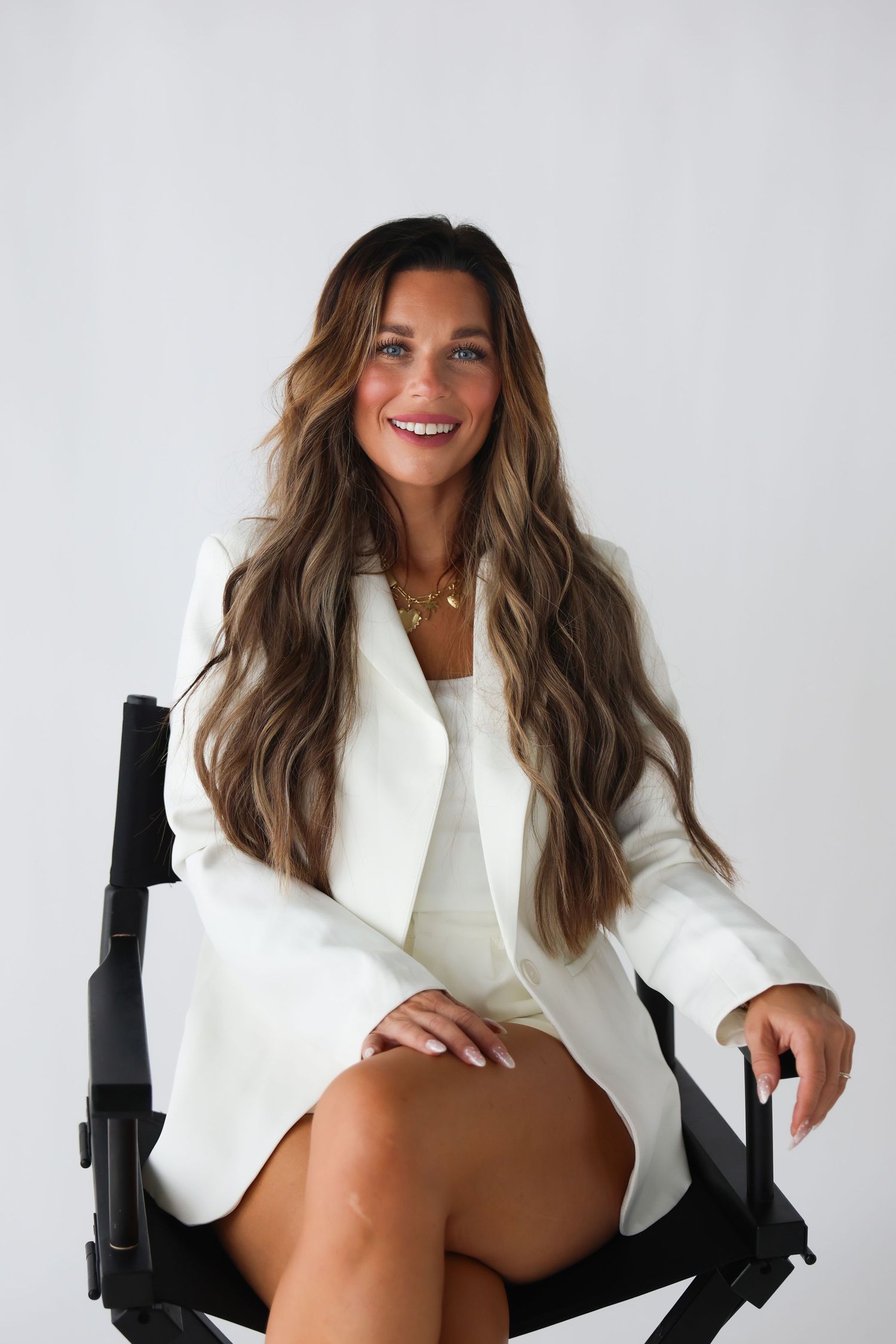Woman in white blazer and dress sitting in black chair, smiling with long wavy brunette hair.