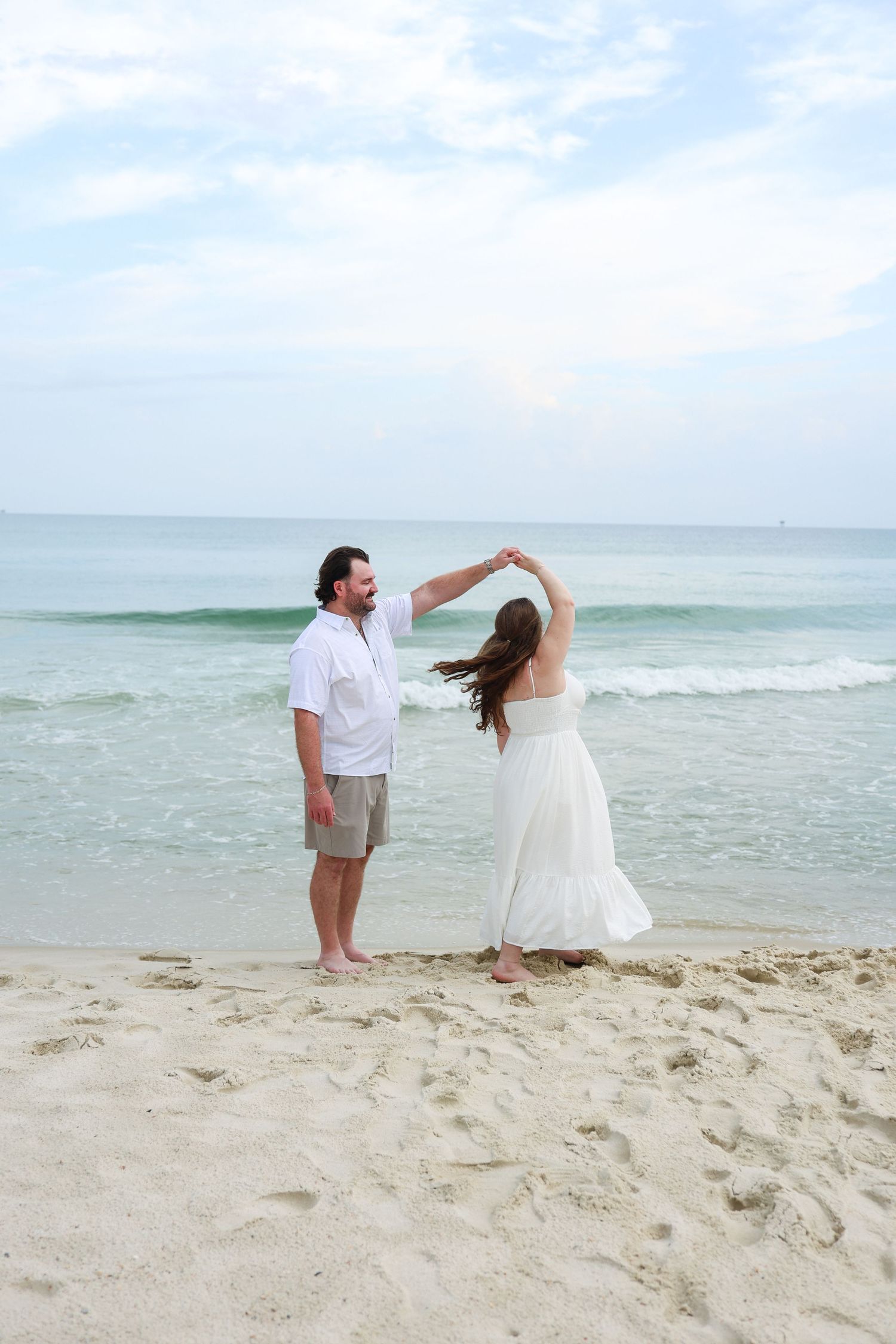 Couple dancing on a sandy beach, man in white shirt and woman in flowing white dress, with ocean waves in the background.