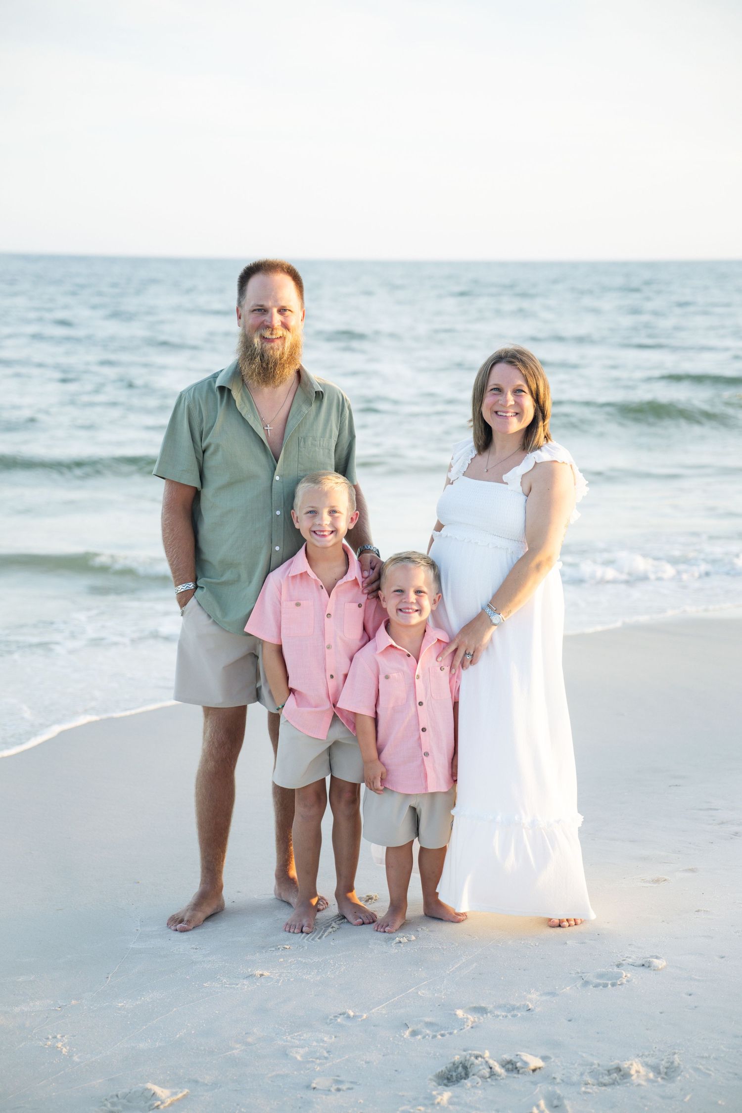 Family of four poses on a sandy beach, parents and two boys in pink shirts, mother in white dress showing baby bump.
