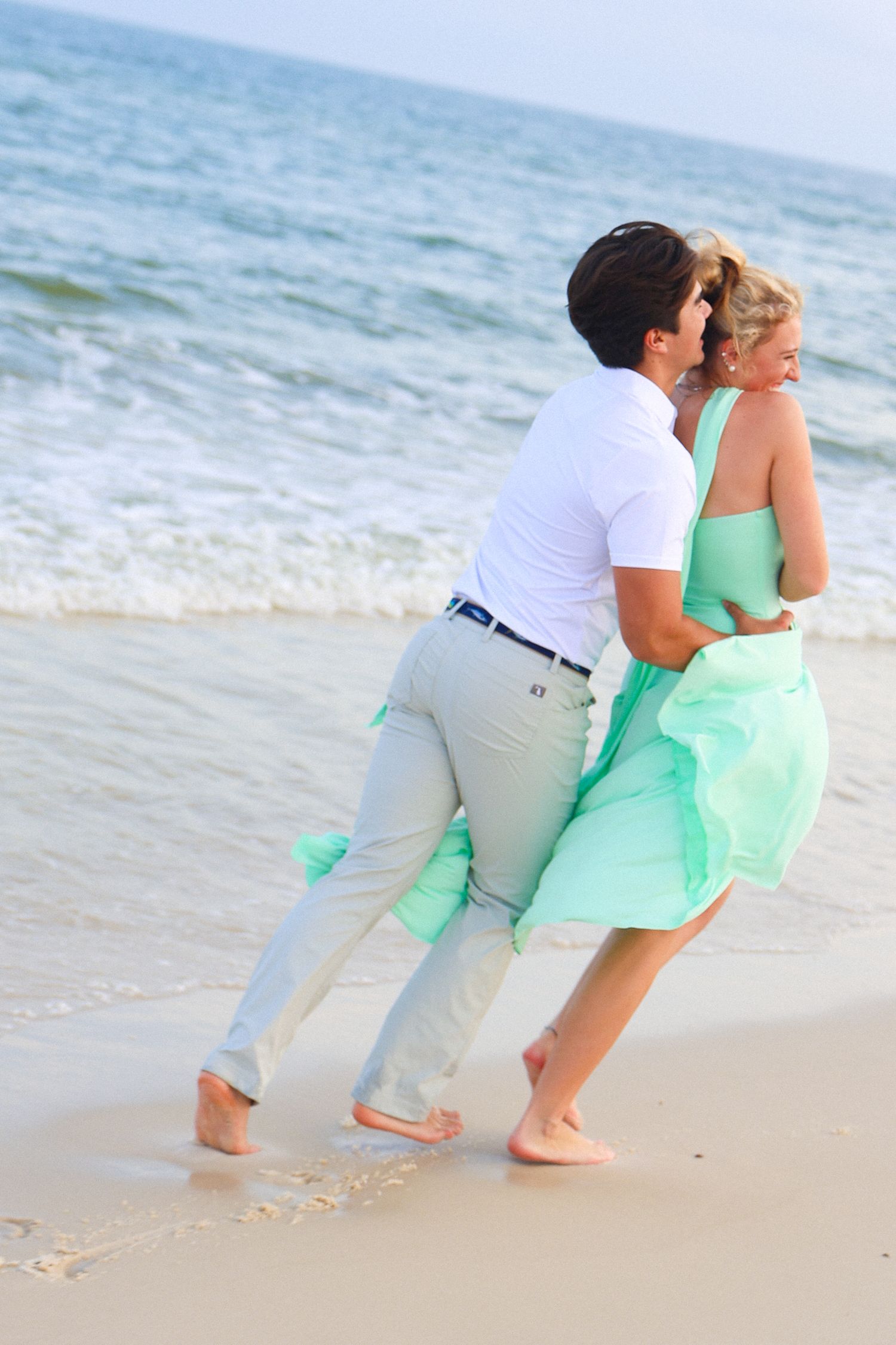 Romantic couple dancing barefoot on a sandy beach, woman in mint green dress, waves in background.