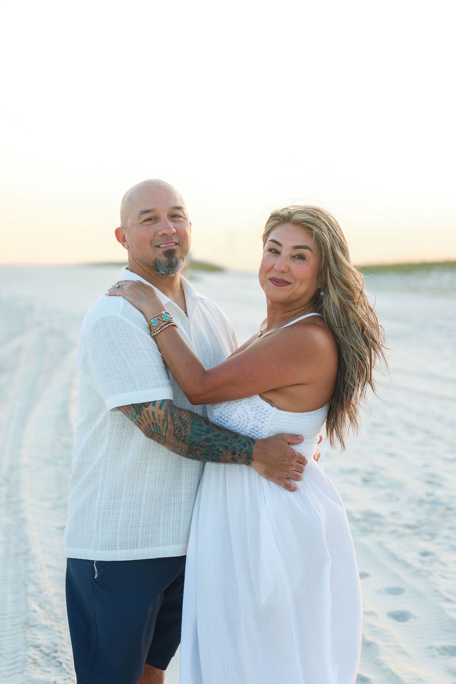 Couple embracing on sandy beach at sunset, man with tattoos in white shirt, woman in white dress.