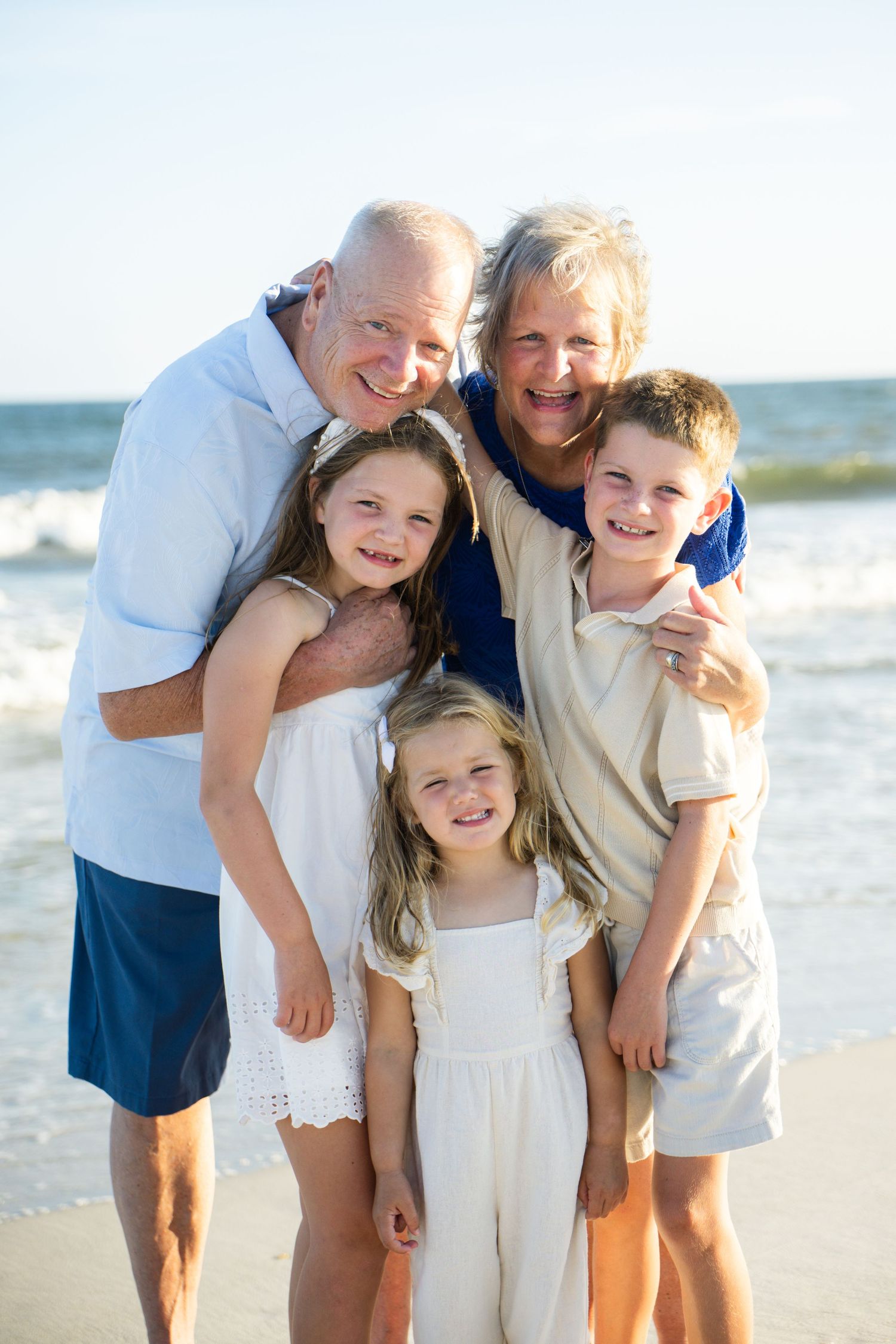 Grandparents smiling with three grandchildren on a sunny beach, posing together for a family portrait.
