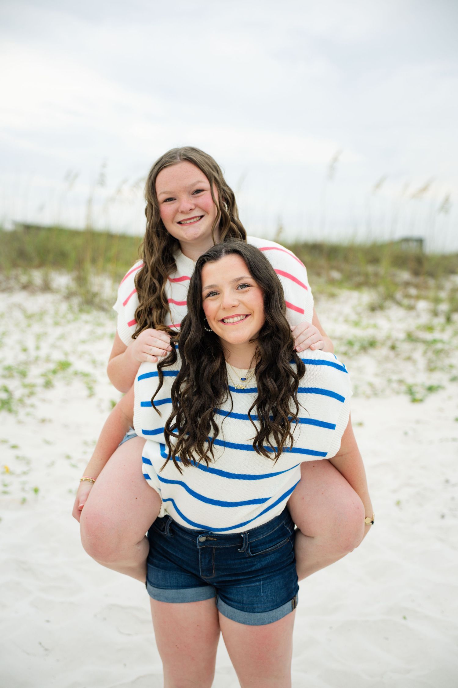 Two smiling girls on a sandy beach, one giving the other a piggyback ride, wearing striped shirts.