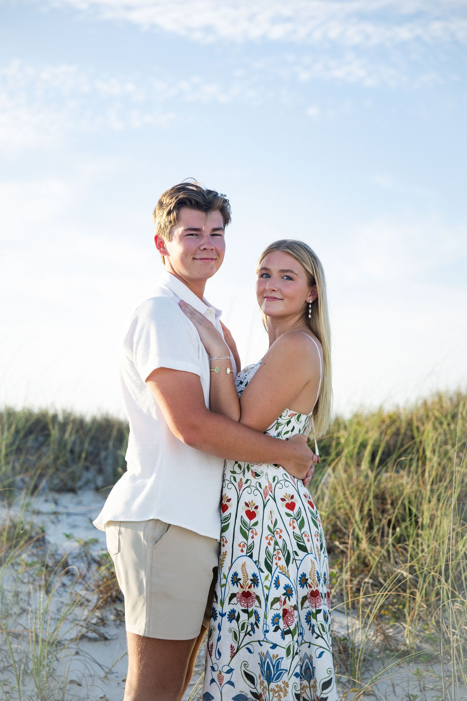 Young couple embracing on sandy beach dunes, woman in floral dress and man in white shirt, blue sky background.