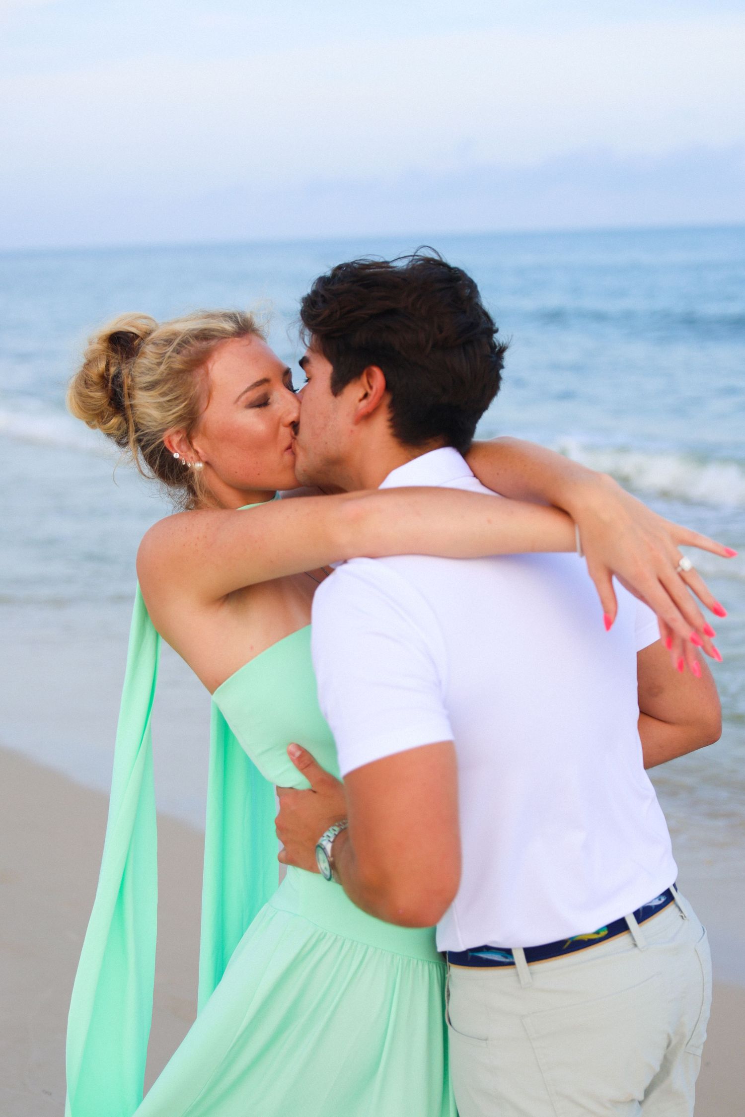 Couple sharing a romantic kiss on a beach, woman in mint green dress, man in white shirt, ocean backdrop.