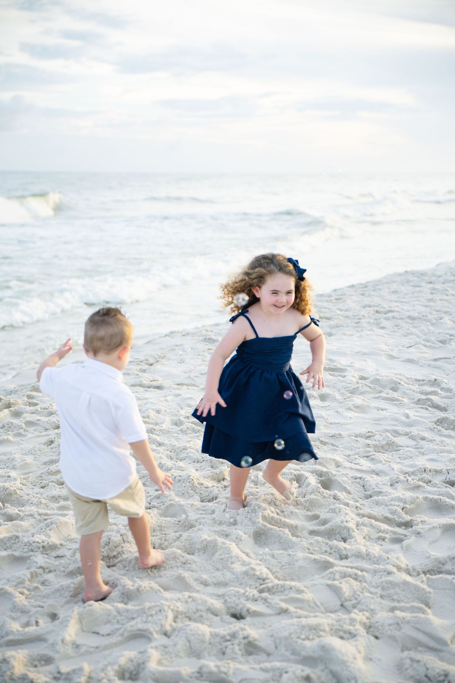 Two young children playing joyfully on a sandy beach, girl in navy dress and boy in white shirt, with ocean waves behind them.