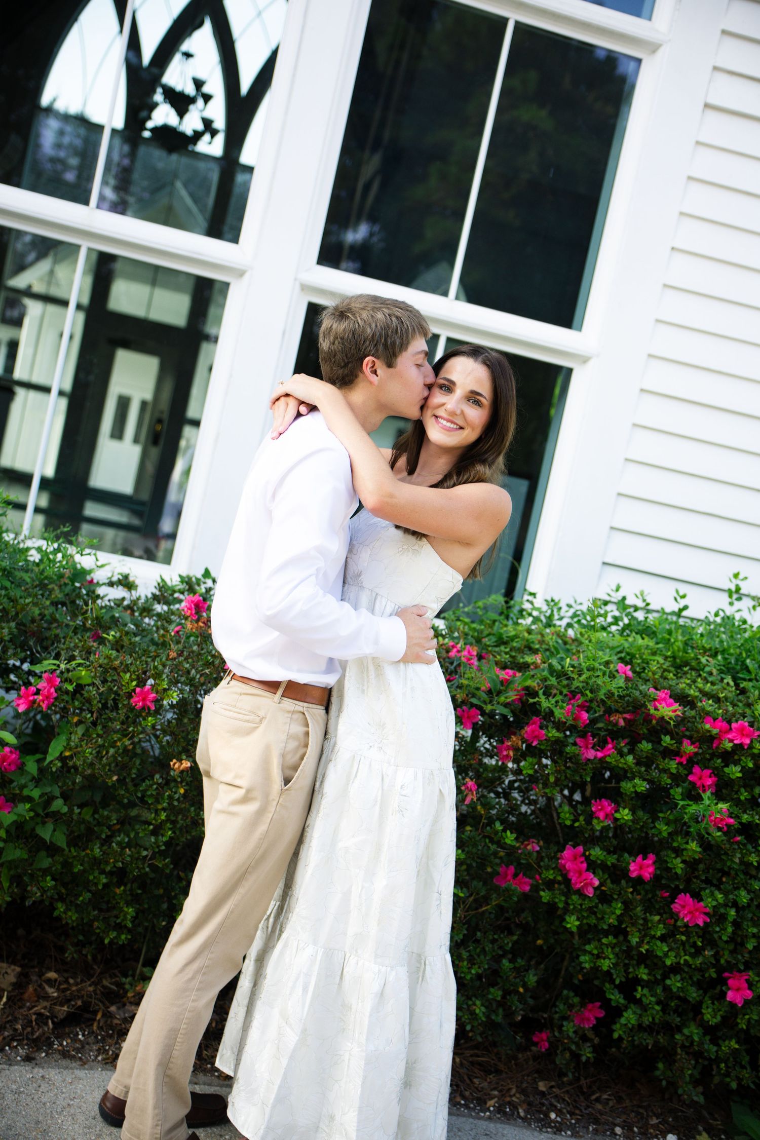 A couple embraces outdoors, man kissing woman's cheek, she smiles in white dress, pink flowers and white building behind them.