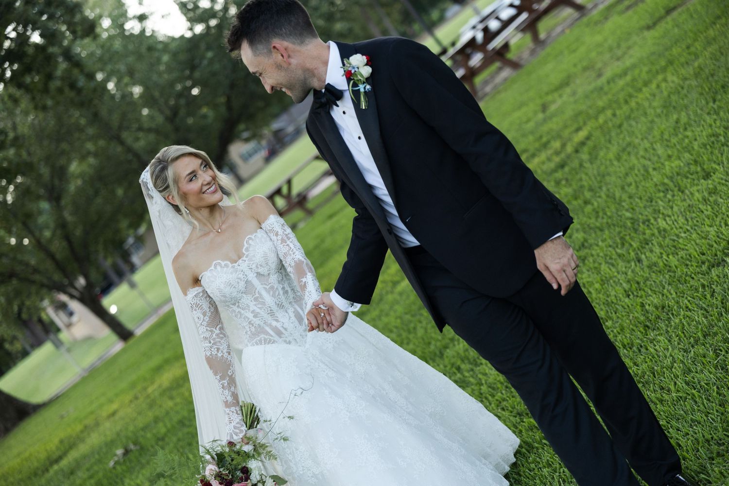 Bride in lace gown and groom in black tuxedo hold hands smiling in a lush green park on their wedding day.