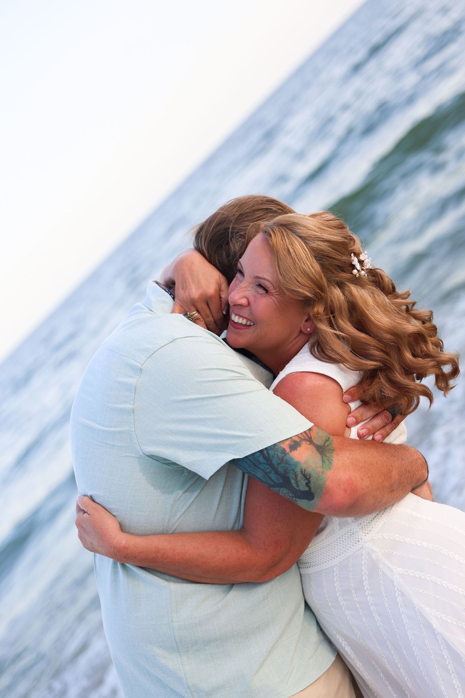 A smiling woman with curly hair hugs a man near the ocean, showcasing a tattoo on her arm.
