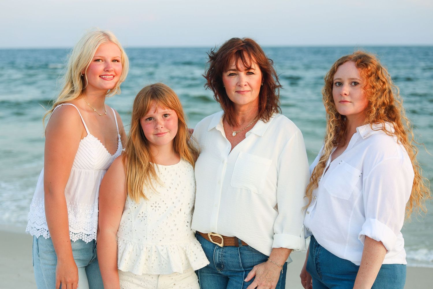 Four women and girls pose together on a sunny beach, wearing white tops and jeans, smiling at the camera.
