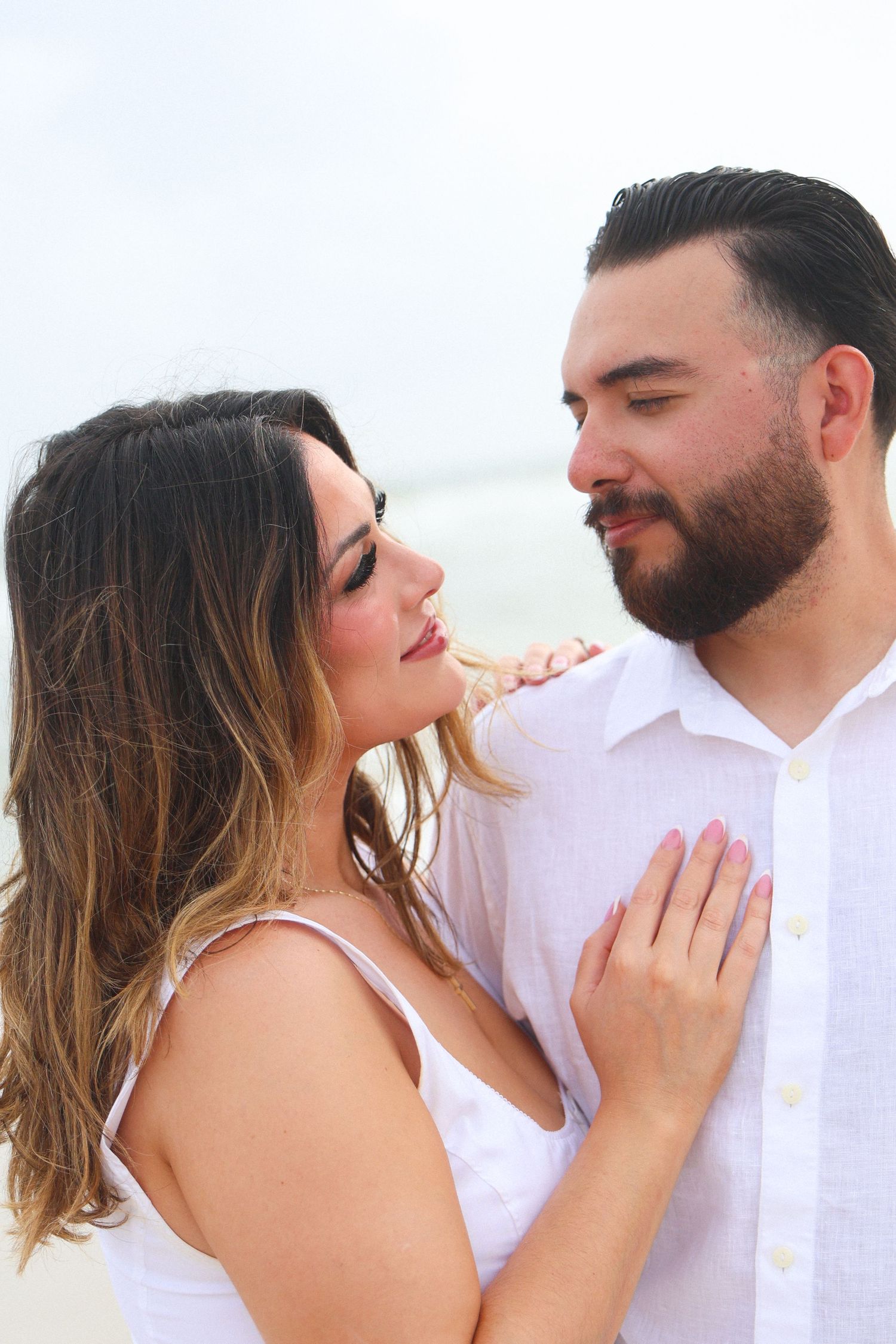 A romantic couple in white outfits share an intimate moment on a beach, the woman gazing up at the bearded man.