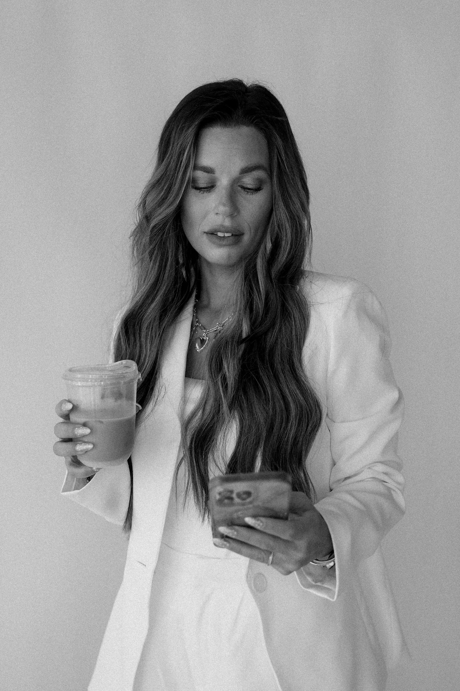 Woman in white blazer holding iced drink and phone, long wavy hair, layered necklaces, black and white photo.