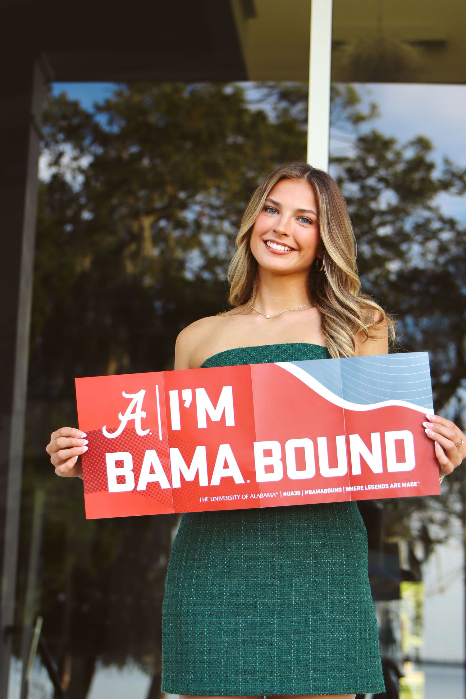 Smiling young woman in green dress holding red University of Alabama 