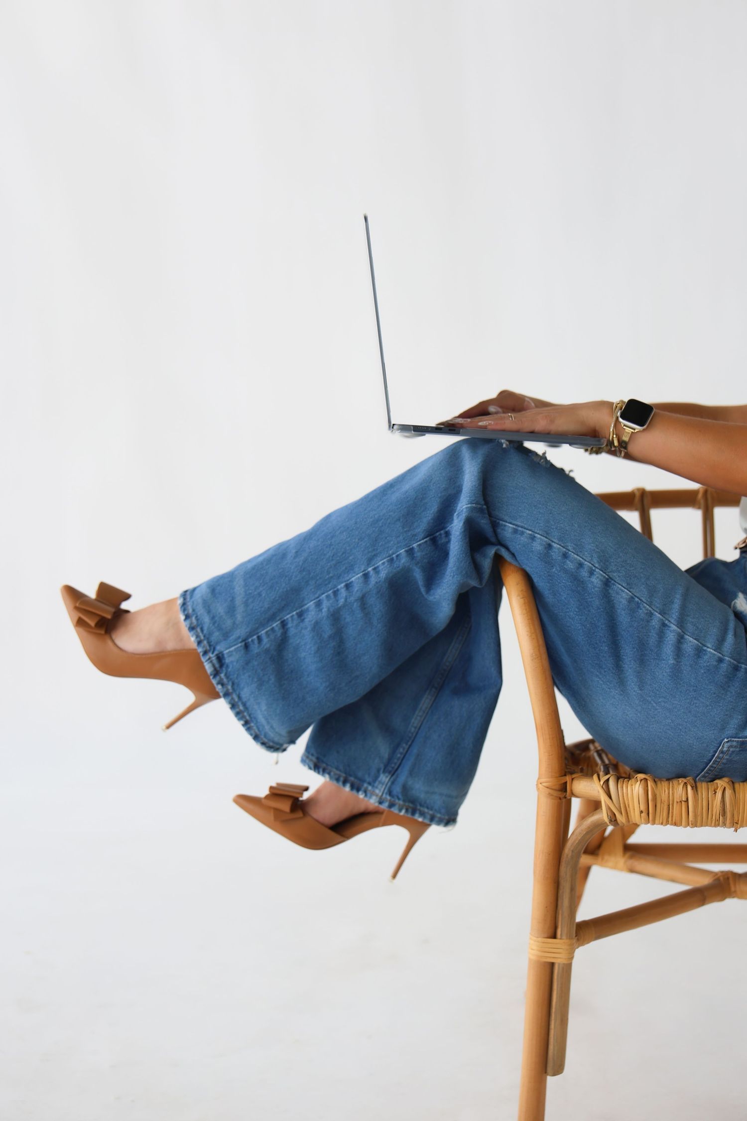 Woman in wide-leg jeans and tan heels sitting in rattan chair with laptop on her lap.