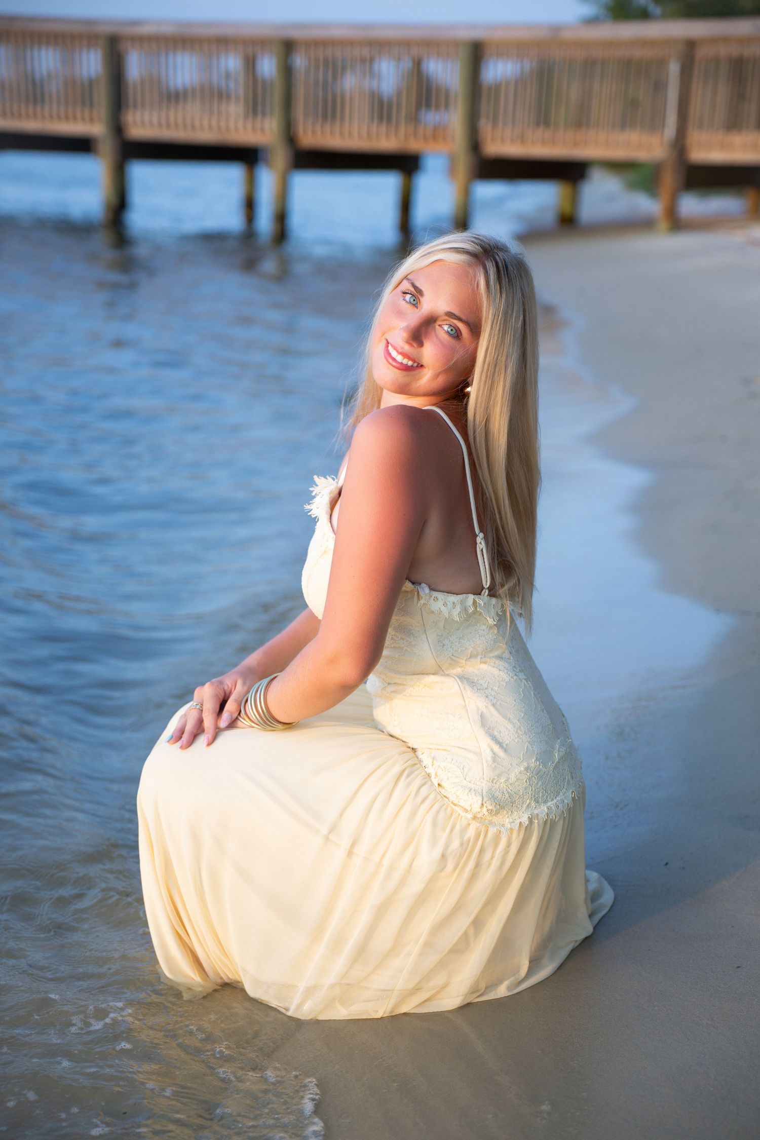Smiling blonde woman in yellow maxi dress sitting on sandy beach shore with wooden pier in background.