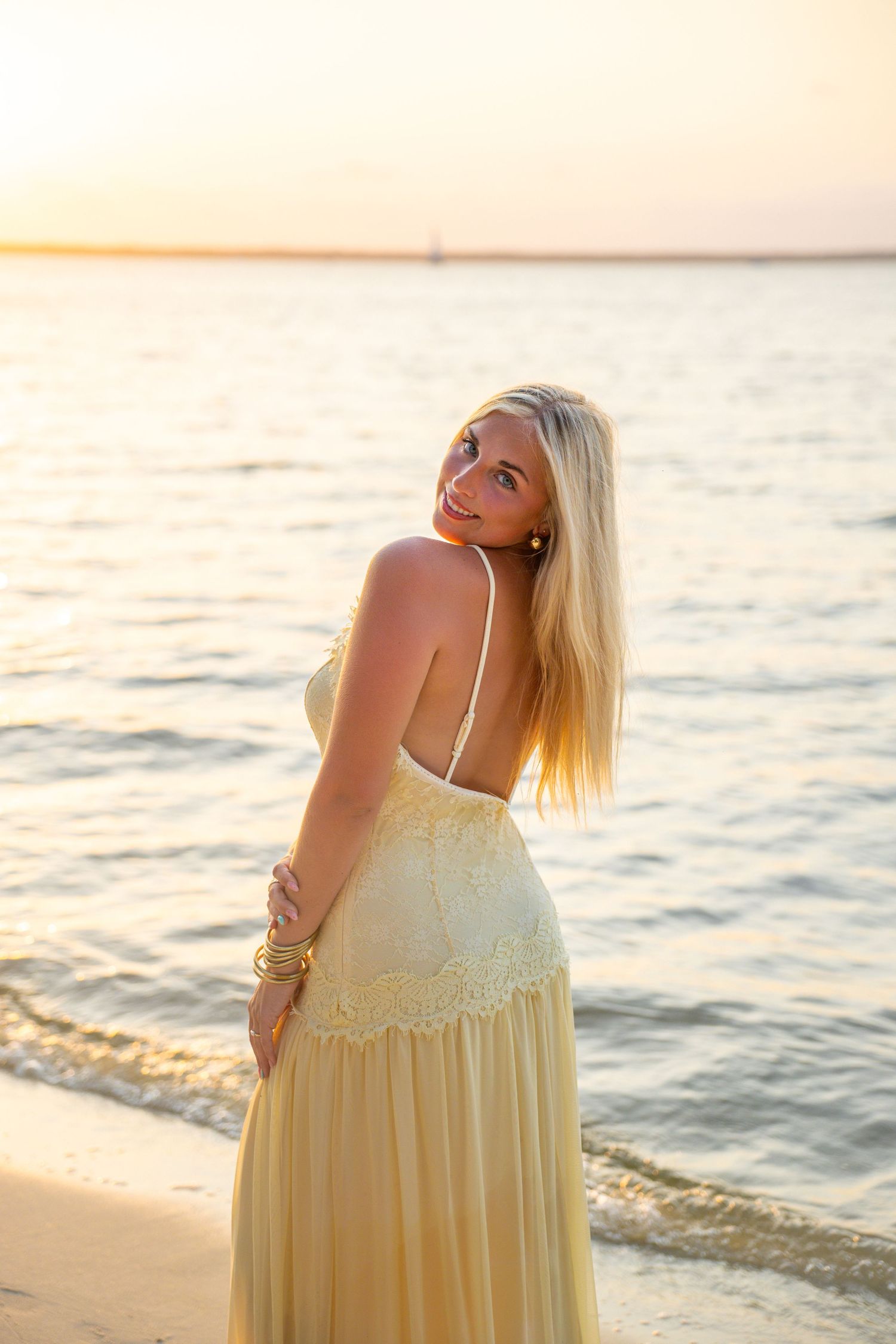 Blonde woman in yellow maxi dress poses on sandy beach at golden sunset with calm water in background.