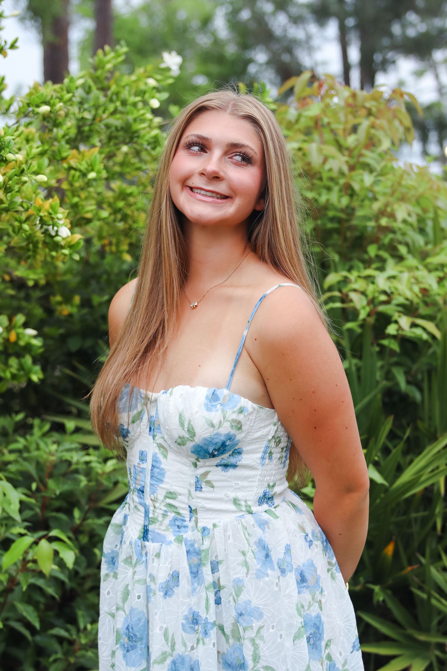 Young woman in a white and blue floral sundress smiling outdoors among lush green bushes.