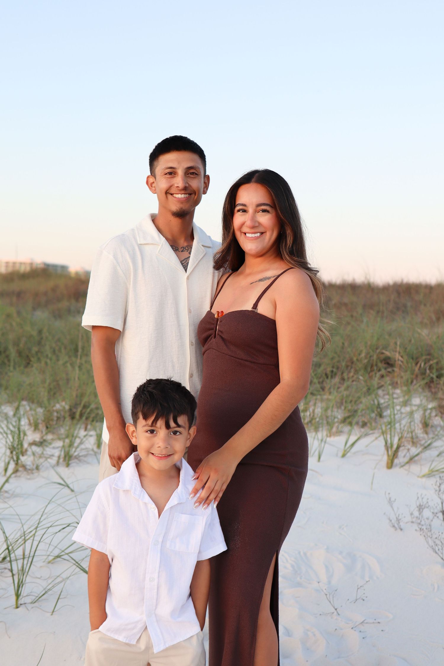 Smiling family of three poses on sandy beach dunes at sunset, pregnant mother in brown dress with partner and young son.