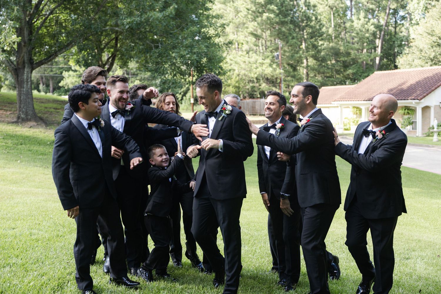 Groom surrounded by groomsmen in black tuxedos laughing and celebrating outdoors on a green lawn at a wedding.