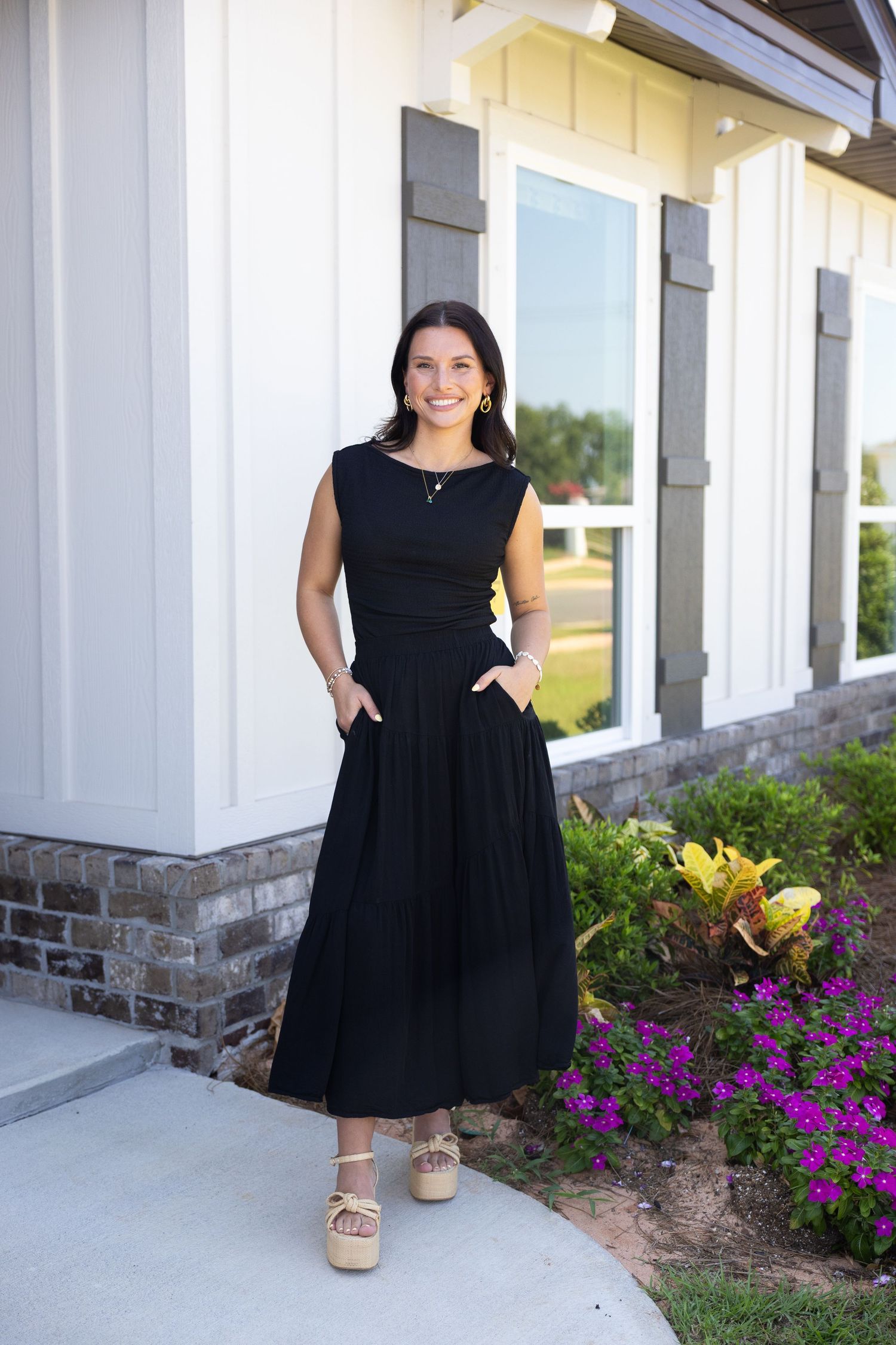 Woman in black sleeveless midi dress with pockets and wedge sandals, smiling outdoors near a building with flowers.