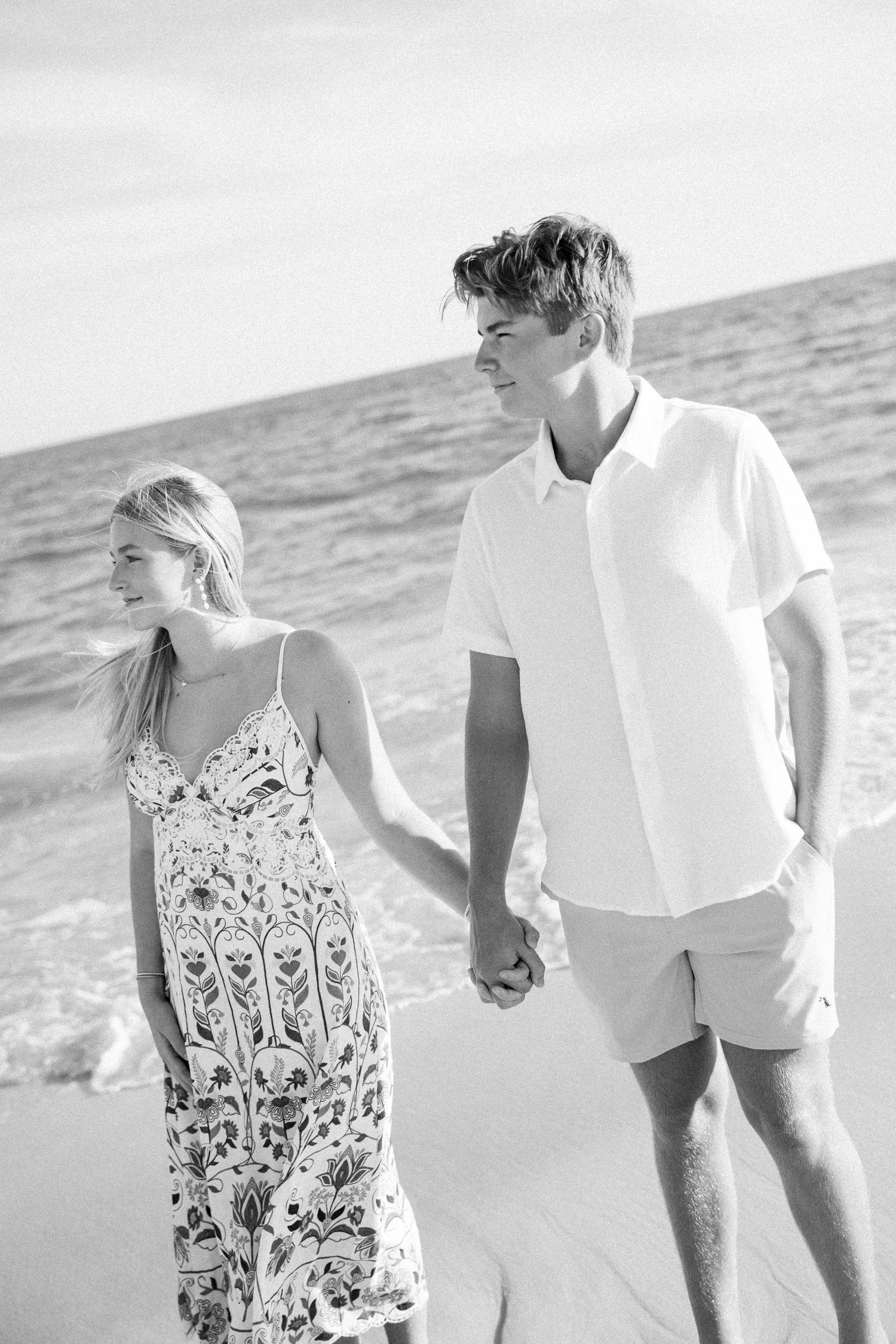 A young couple holds hands while walking on a beach, the man in a white shirt and the woman in a floral sundress.