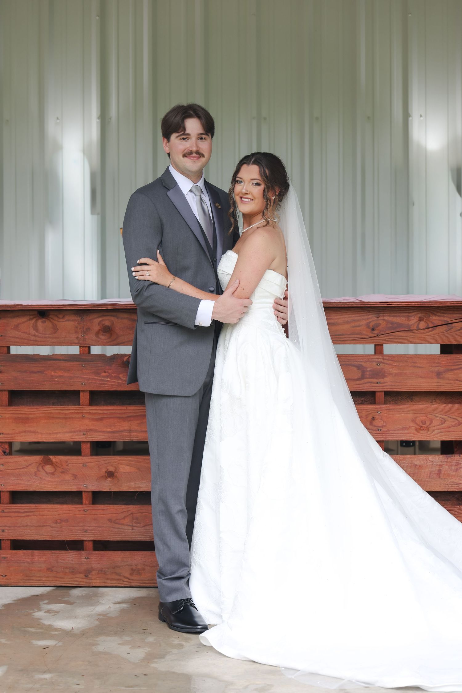 A bride in a white gown and veil poses with a groom in a gray suit at a rustic wooden fence venue.