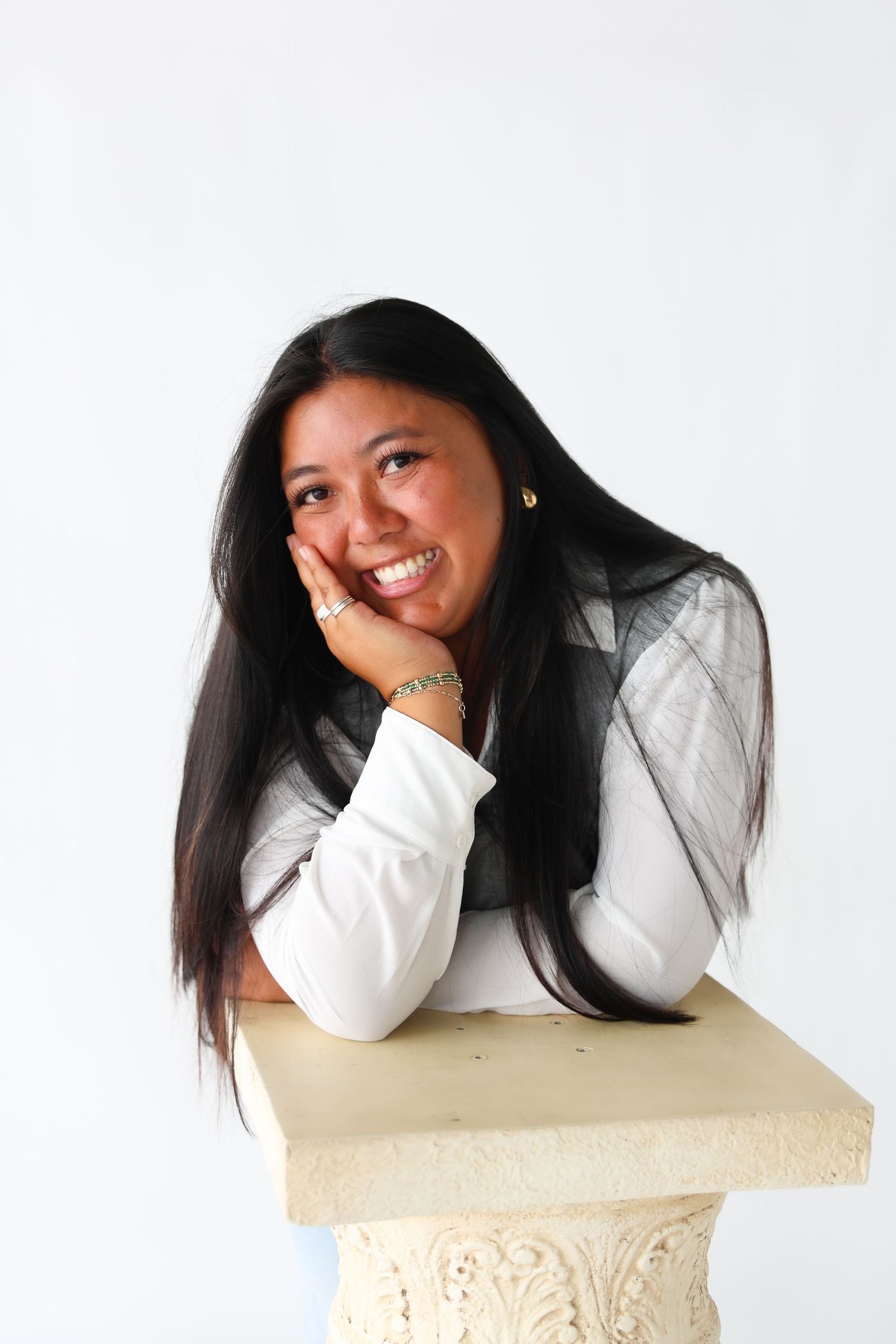 Smiling young woman with long black hair resting her chin on her hand, leaning on a decorative stone pedestal.