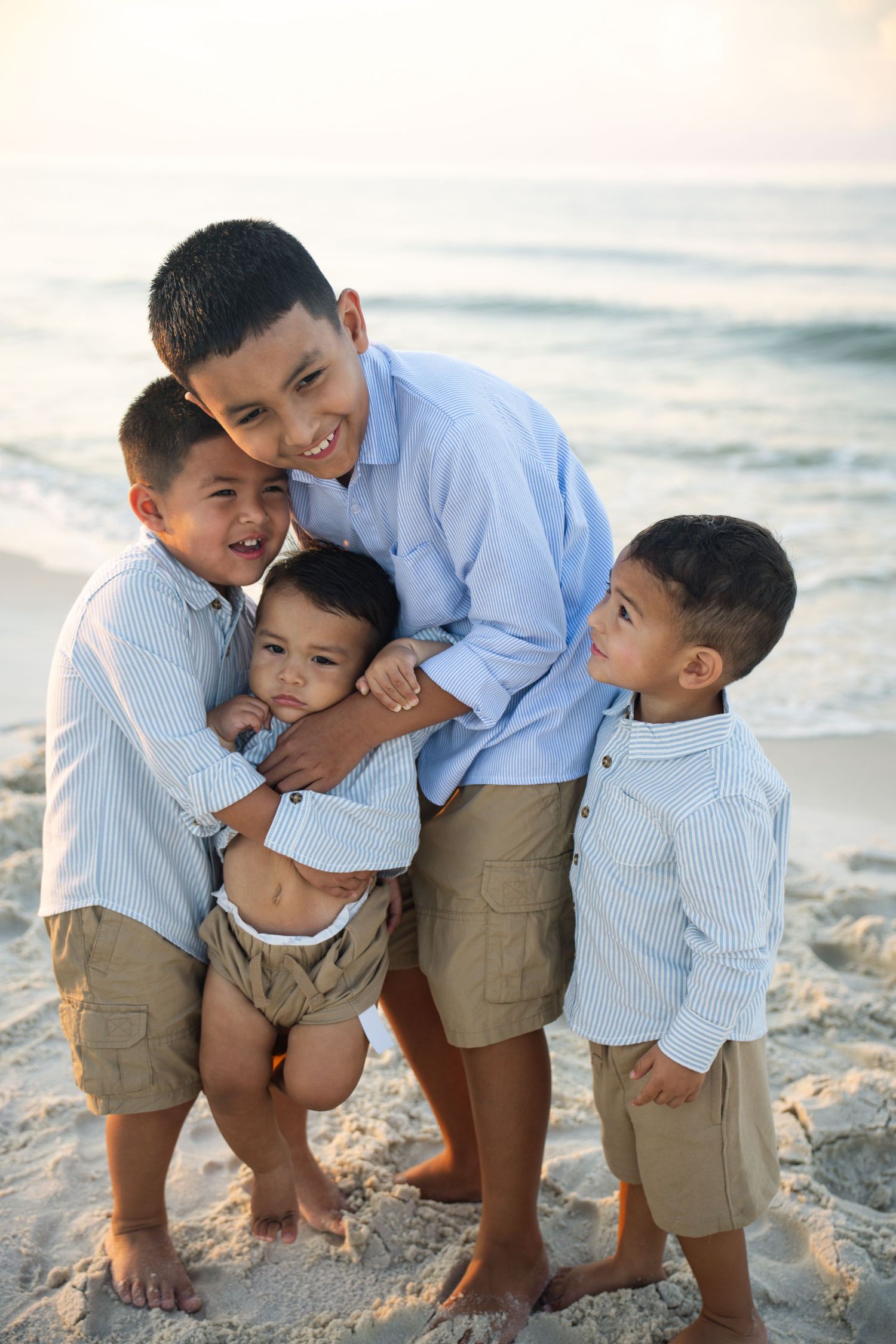 Four young brothers pose together on a sandy beach, smiling and hugging near ocean waves at sunset.