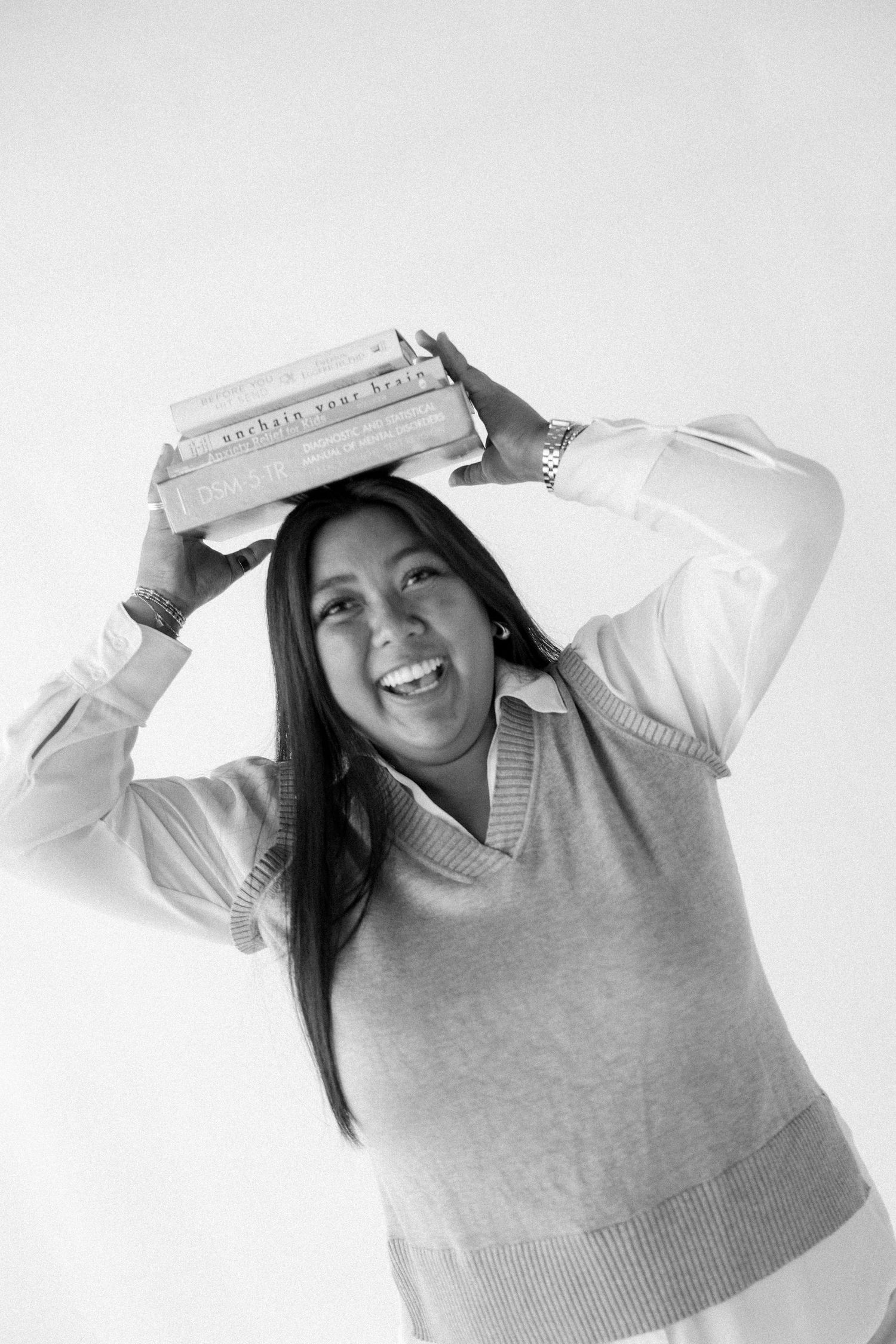 Smiling woman balancing a stack of books on her head, wearing a sweater vest, in a black and white photo.