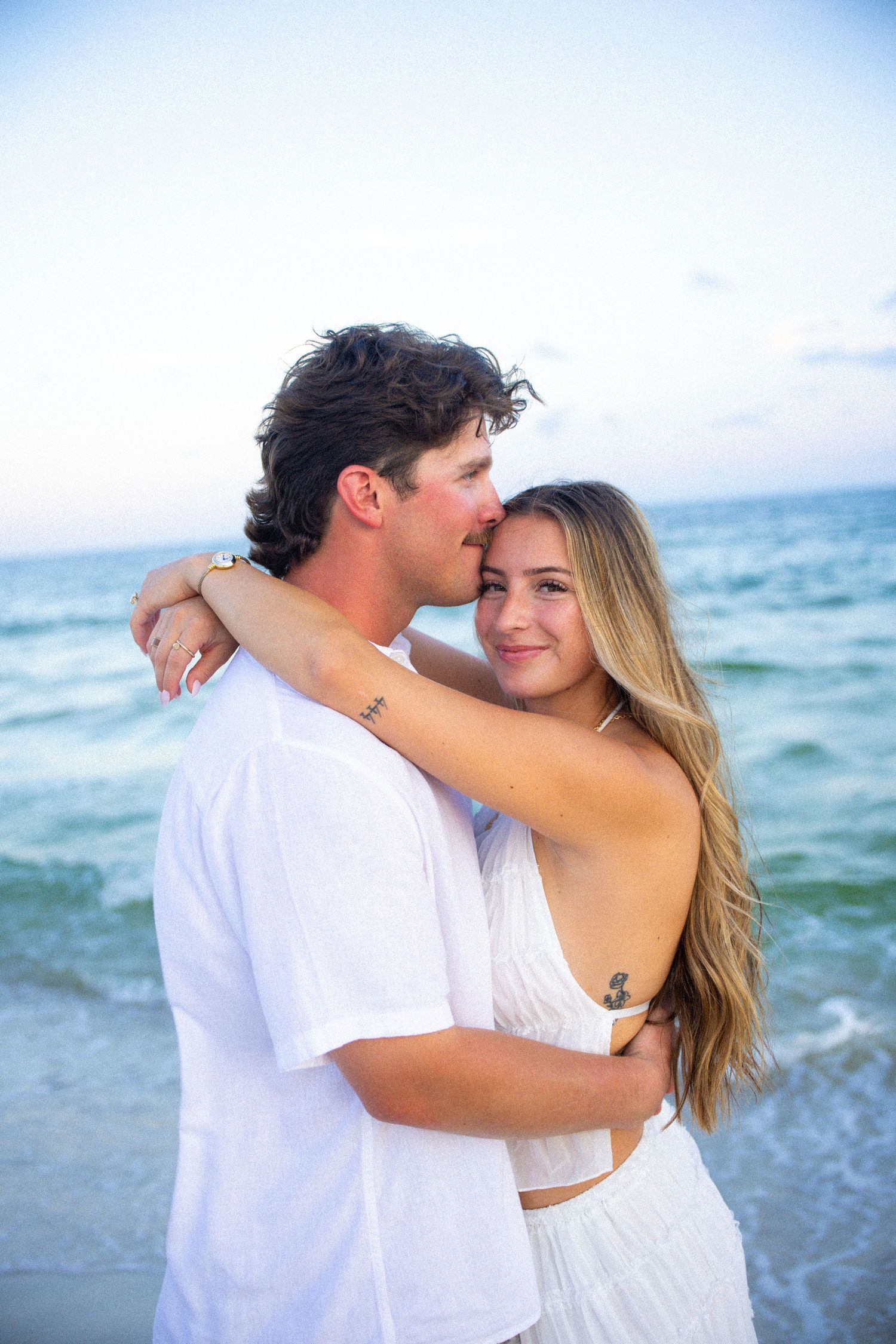 A young couple in white outfits embracing on a beach at dusk, with ocean waves in the background.