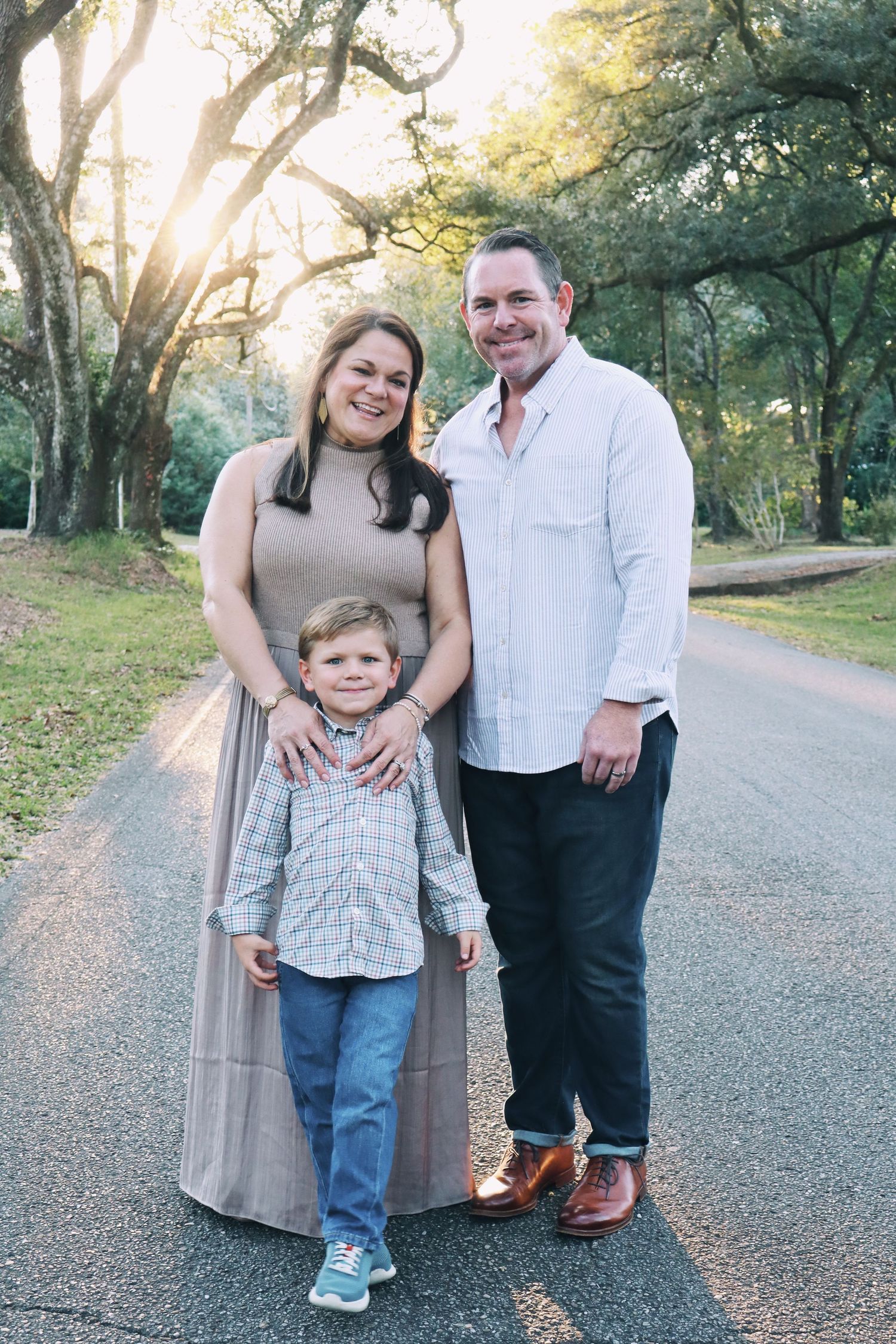 A smiling family of three poses on a tree-lined road at golden hour sunset.