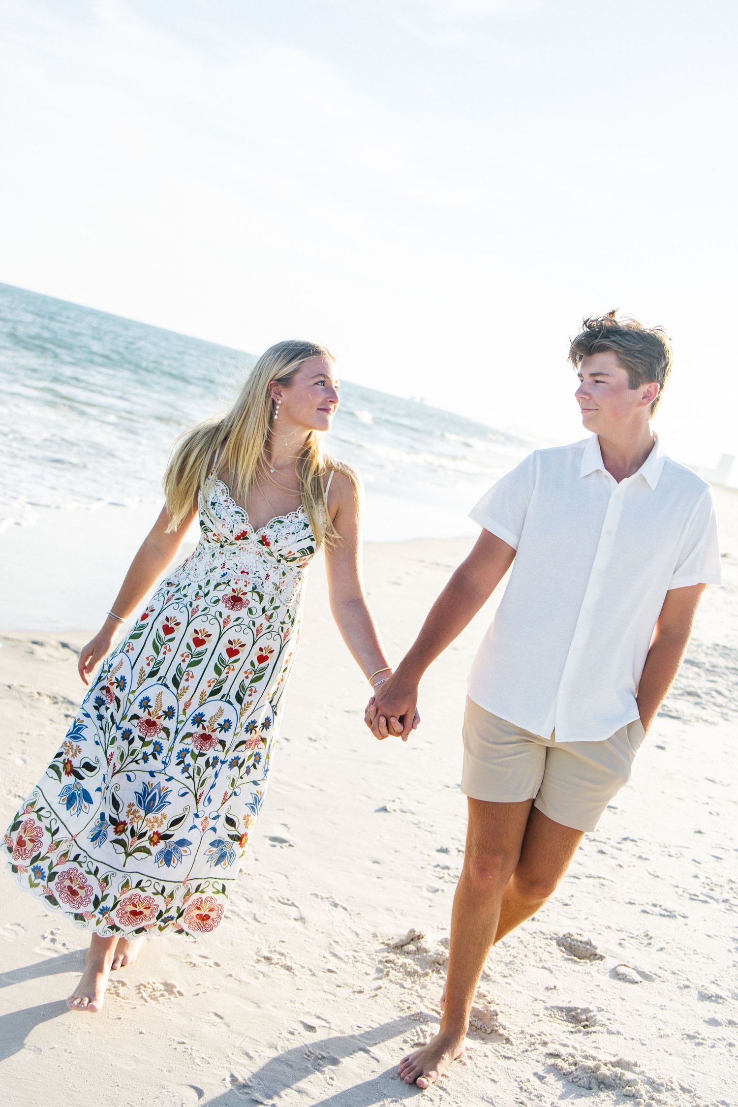 A young couple holds hands and walks along a sandy beach, smiling at each other in bright summer outfits.
