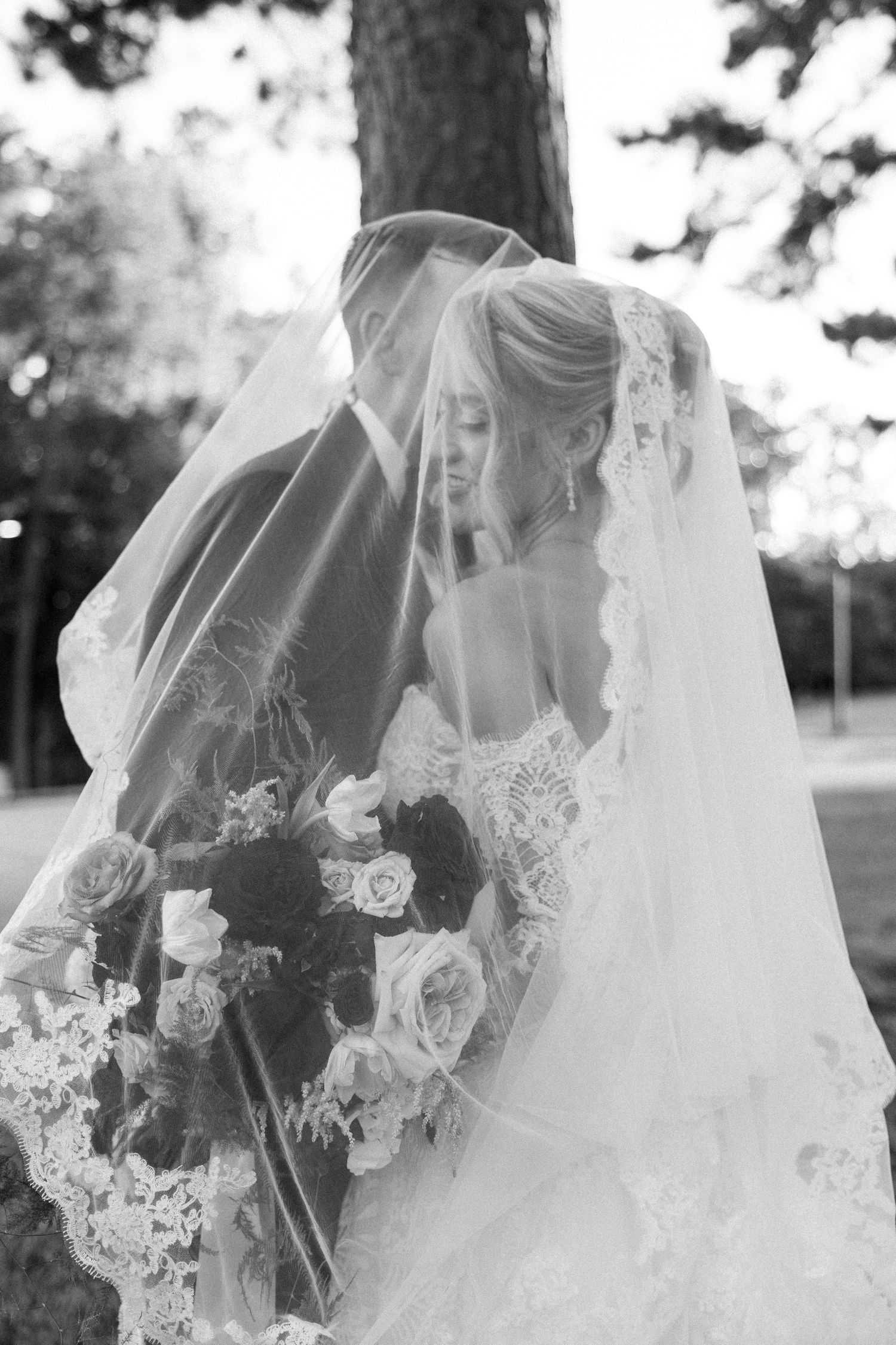 Black and white wedding photo of bride and groom sharing a kiss under lace veil, holding floral bouquet outdoors.