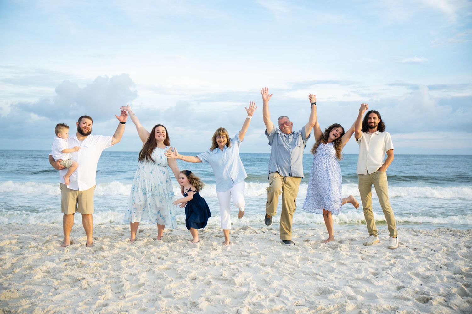 Happy family of seven celebrating on a sunny beach, arms raised joyfully, wearing casual summer outfits with ocean waves behind them.