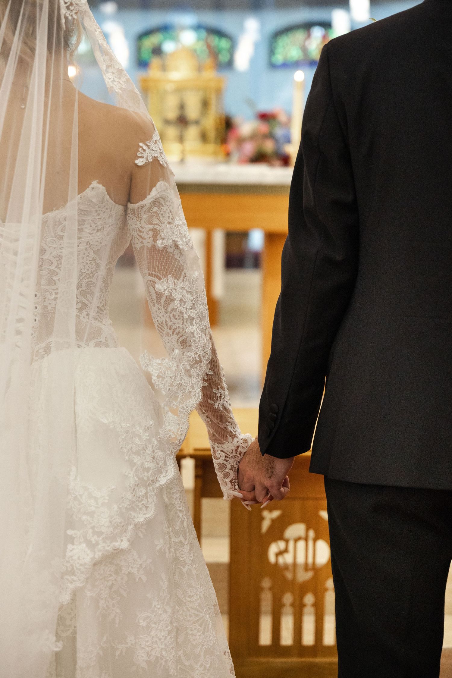 Bride and groom holding hands at church altar, bride wearing lace long-sleeve wedding gown with cathedral veil.