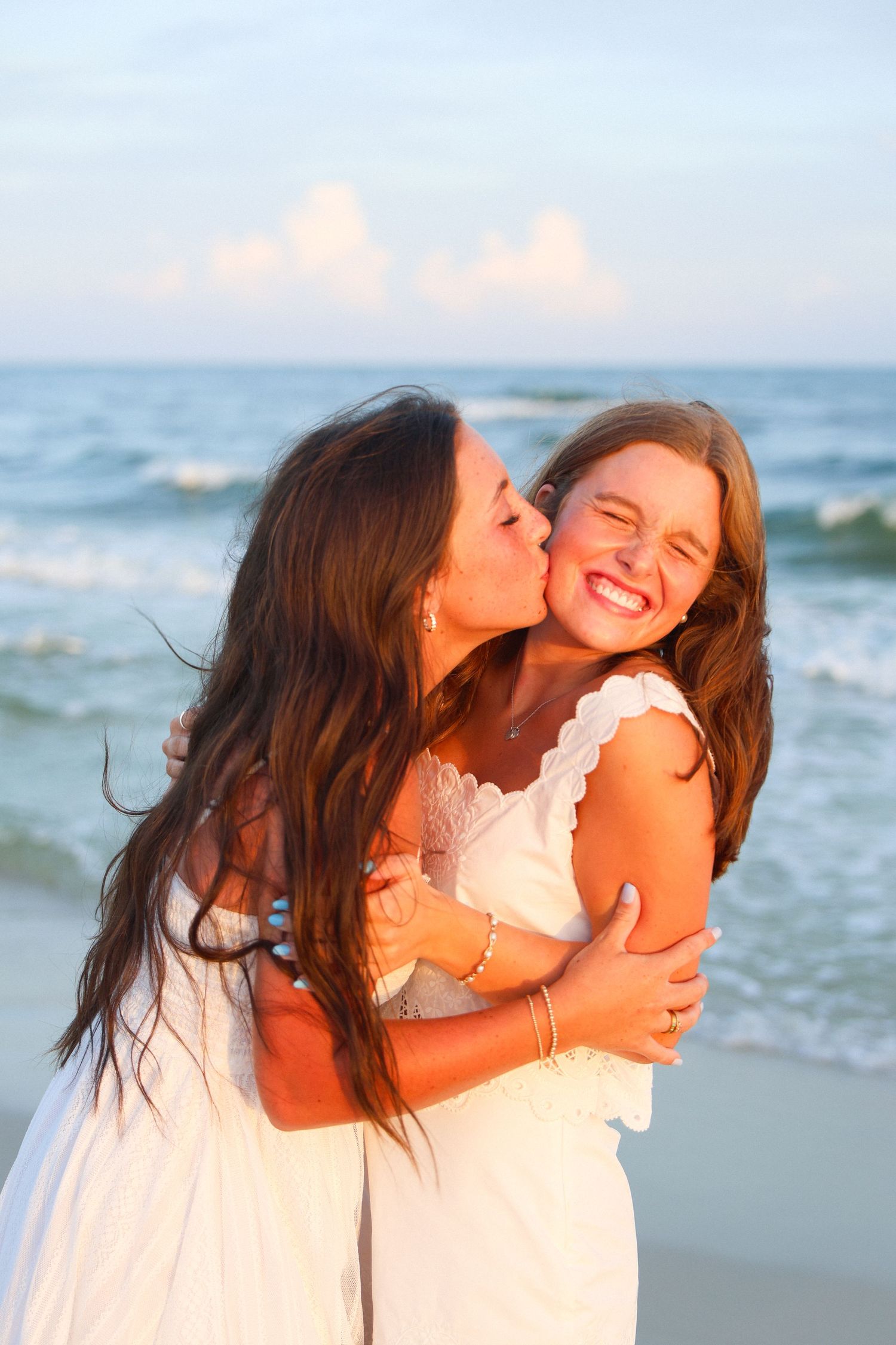 Mother kissing her laughing daughter on the cheek while hugging on a sunny beach, both wearing white dresses.