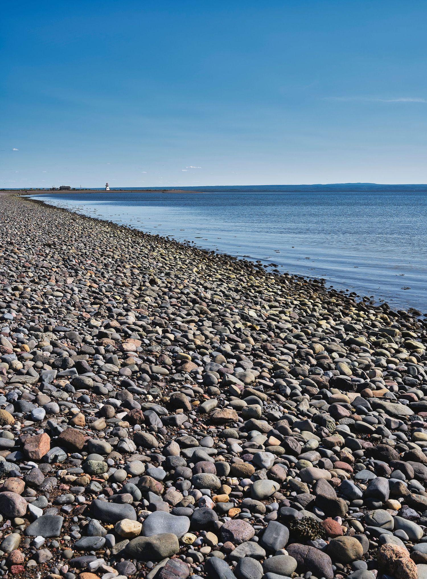 Rocky beach covered in smooth stones stretches along calm ocean waters under clear blue sky.