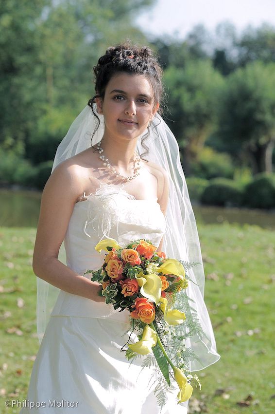 Bride in white strapless gown and veil holds orange rose bouquet outdoors on green lawn.