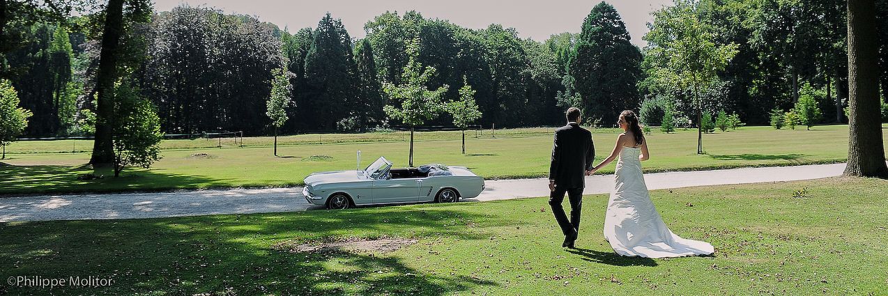 A bride and groom walk hand in hand across a lush green lawn beside a classic white vintage car on their wedding day.