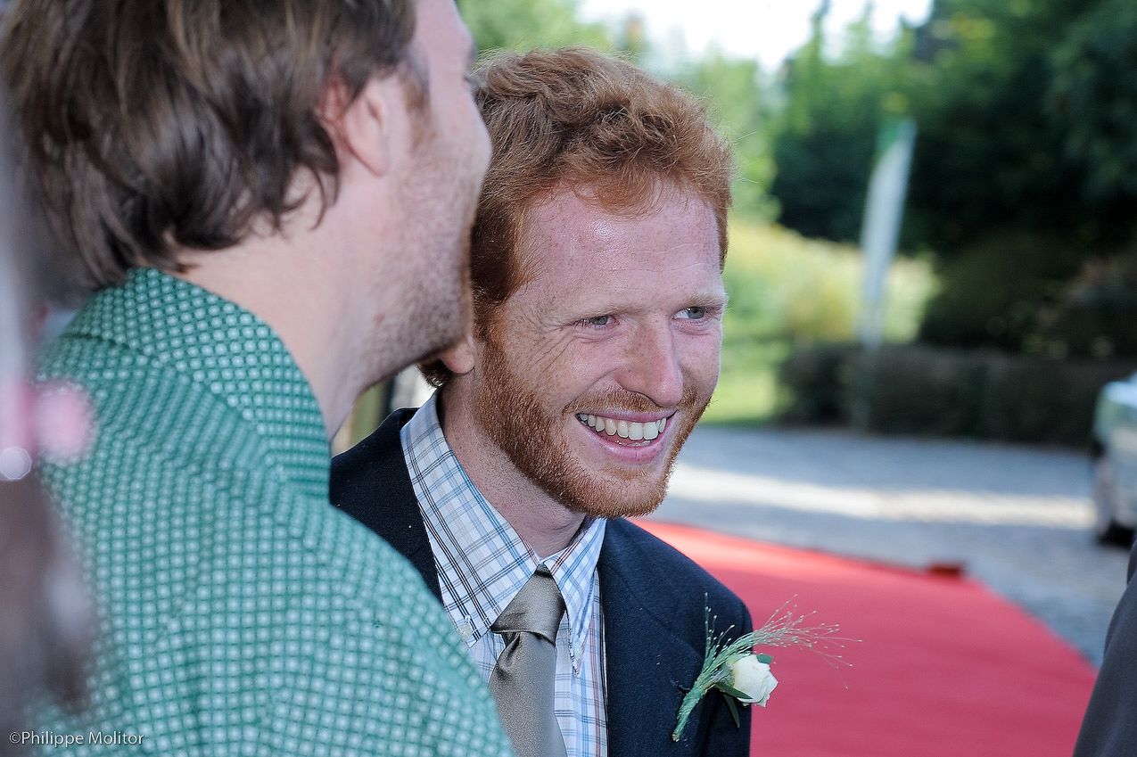 Smiling red-haired man with beard wearing suit and boutonniere at outdoor wedding event.