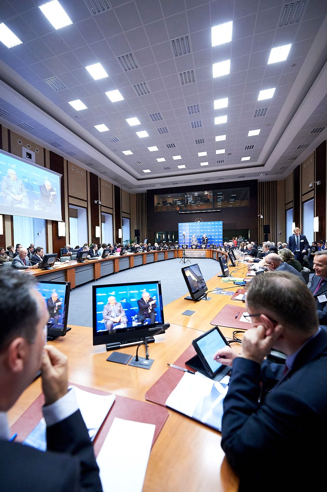 Large conference room with digital displays and attendees seated at curved table arrangement.